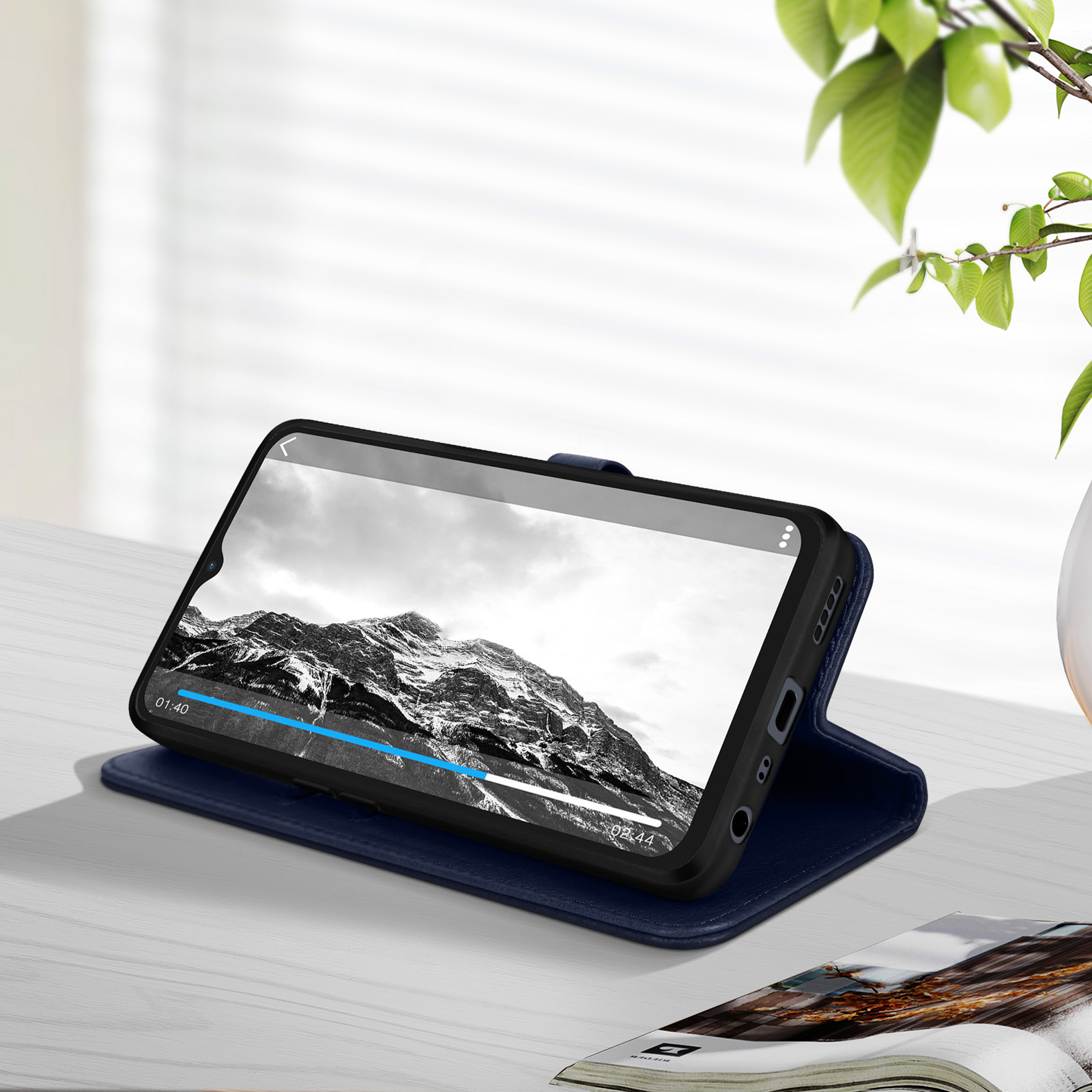 Uno smartphone in una custodia blu mostra un video su un tavolo con una rivista e delle foglie.
