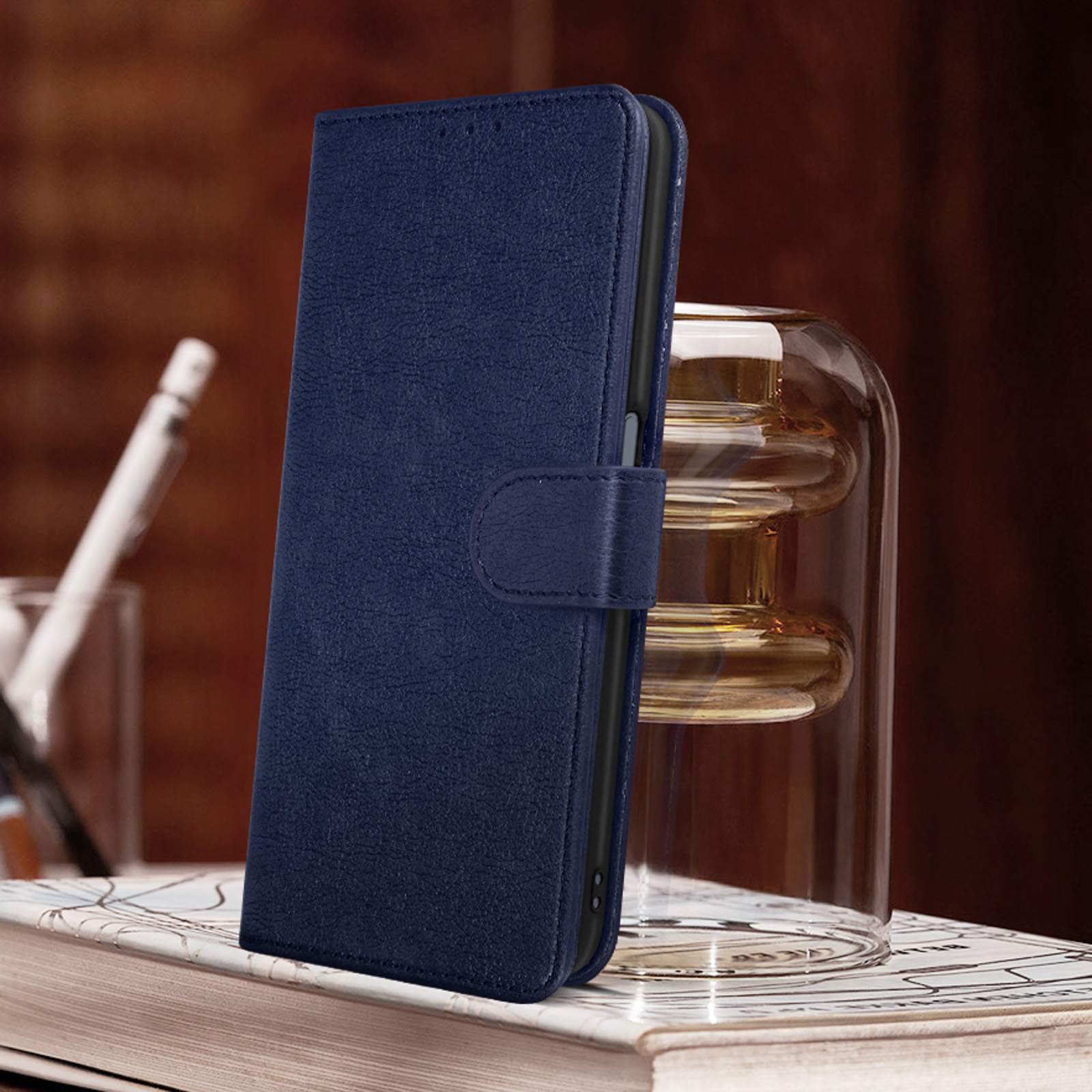 Una custodia blu navy, chiusa e in piedi su una superficie di legno. Un vaso di vetro e una penna dietro.