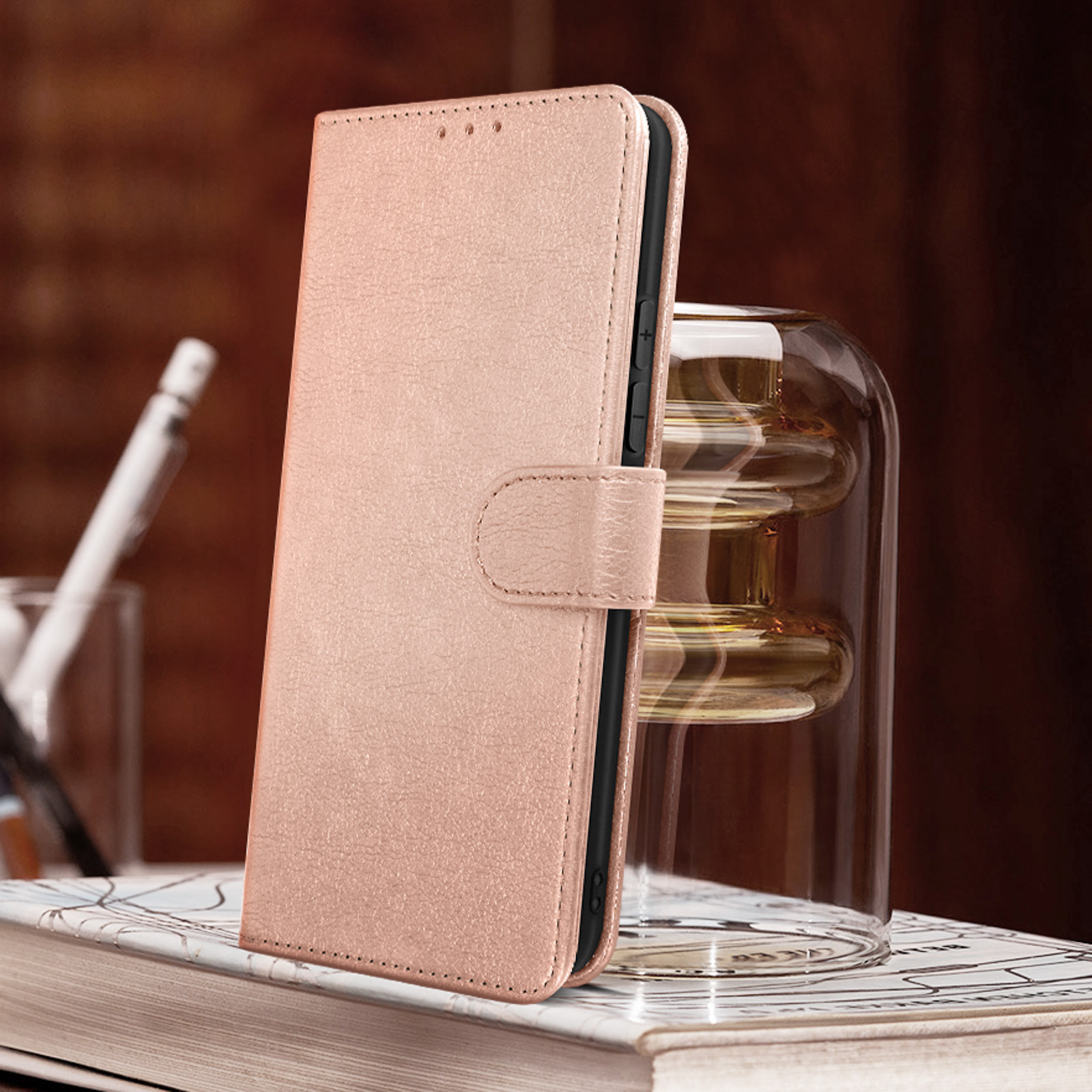 Un étui de téléphone en cuir rose est sur une surface en bois. Il est ouvert, avec un stylo doré et un vase en verre.