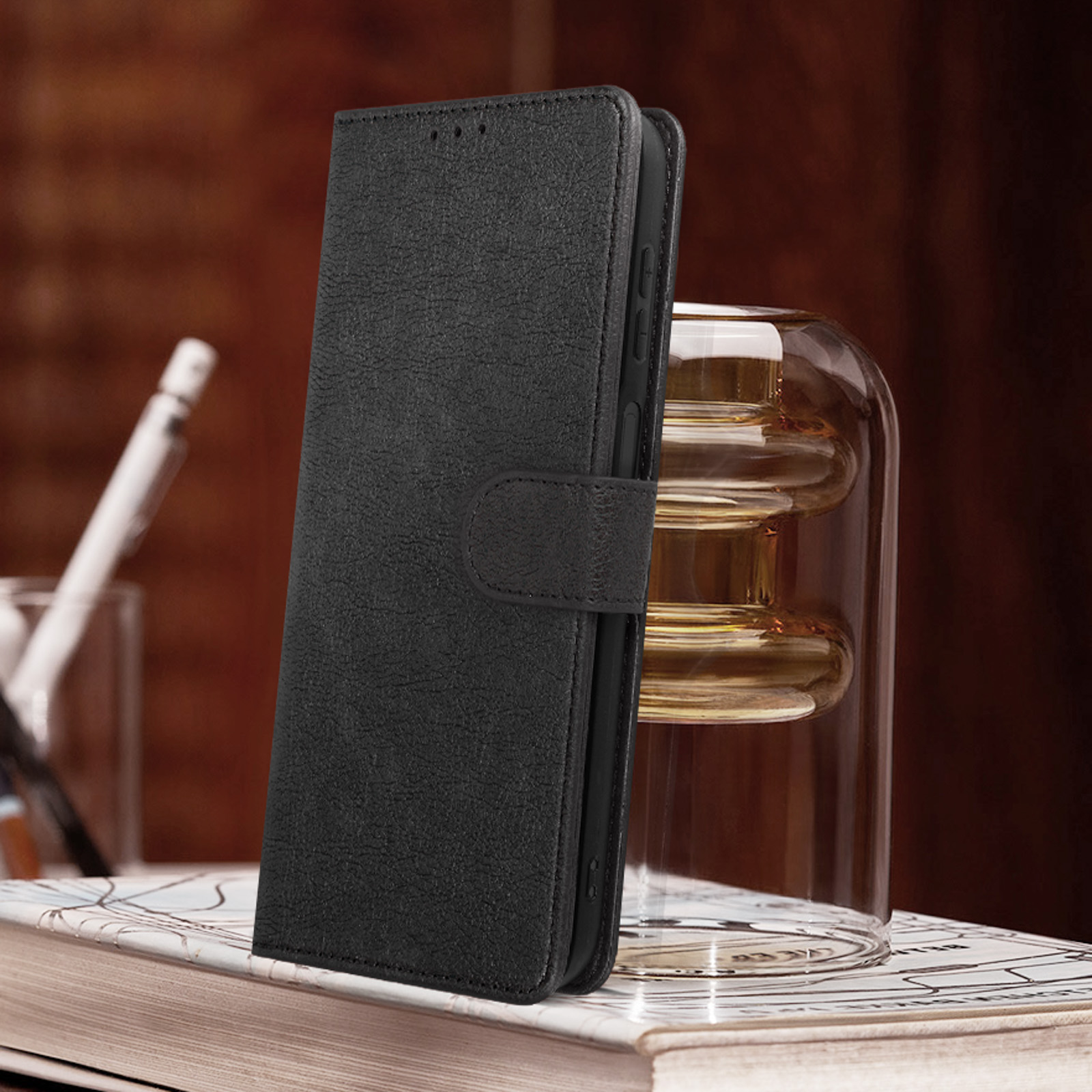 Een zwarte leren telefoonhoes, afgesloten met een bandje. Het staat op een boek naast een glas.