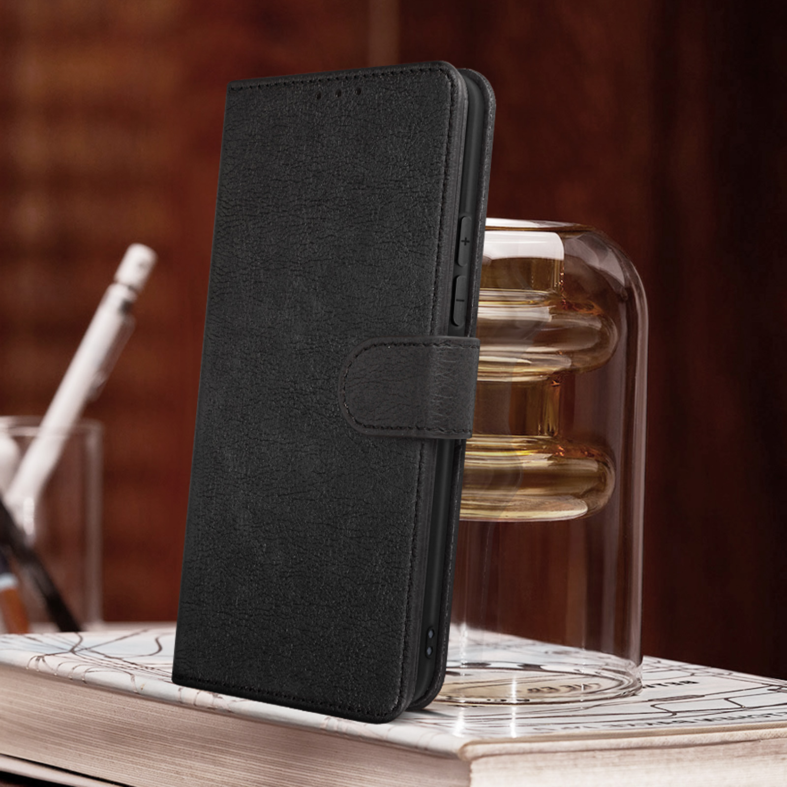 Étui de téléphone en cuir noir, fermé avec une lanière, posé sur des livres à côté d'un récipient en verre.
