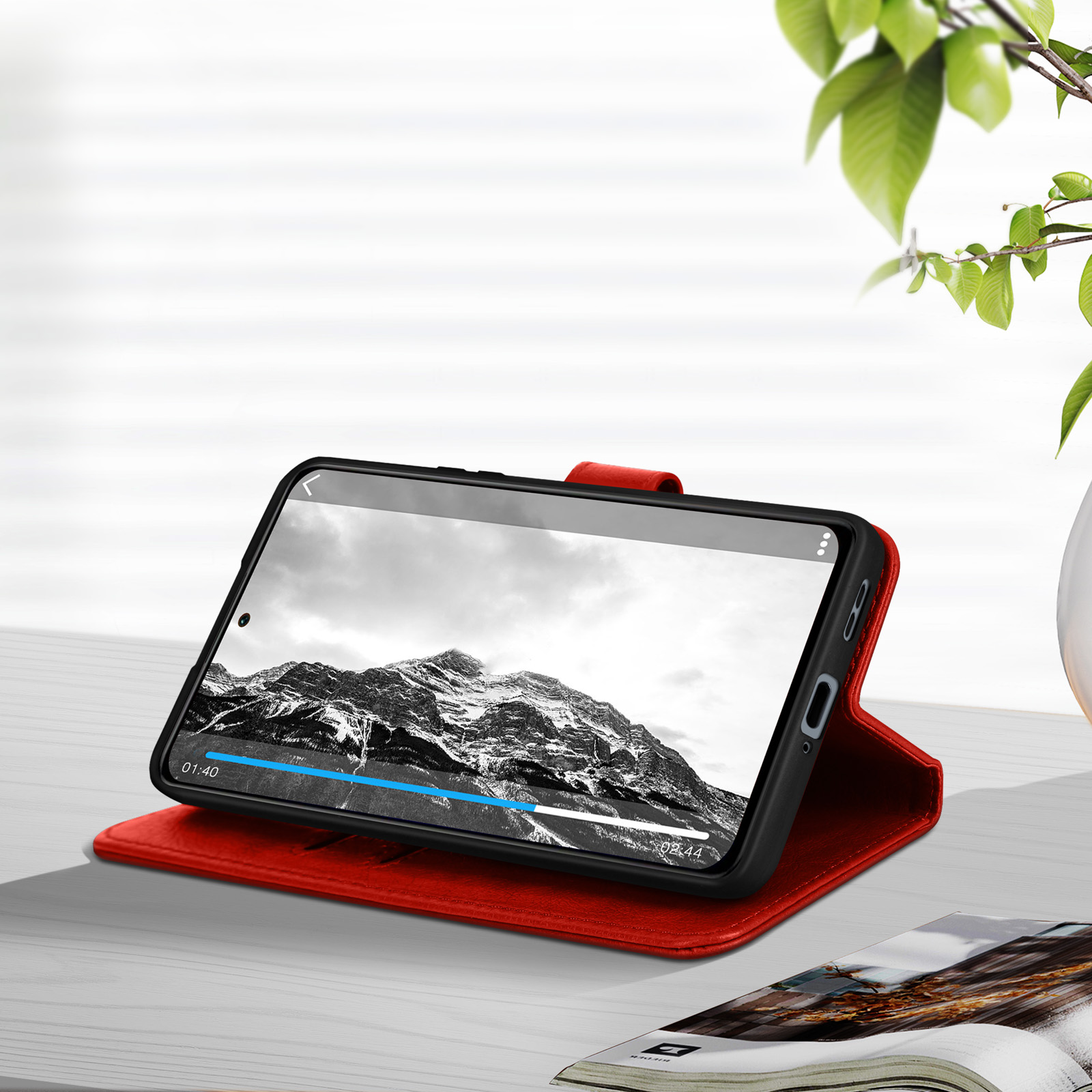 Uno smartphone in una custodia rossa mostra un paesaggio montano su un tavolo con una rivista e delle foglie.