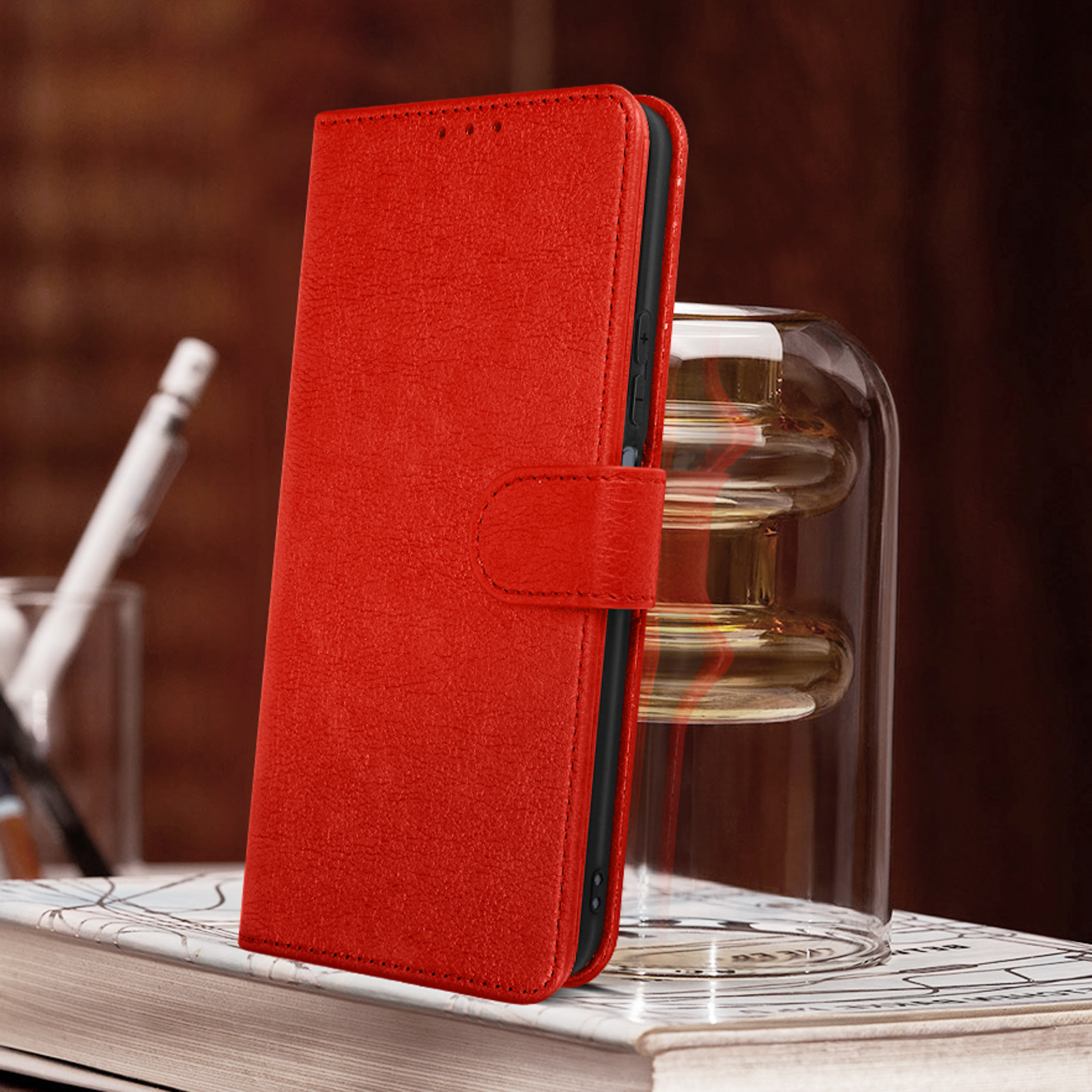 Custodia per telefono rossa, chiusa, su un libro. Vaso di vetro e penna sullo sfondo, sfocati.