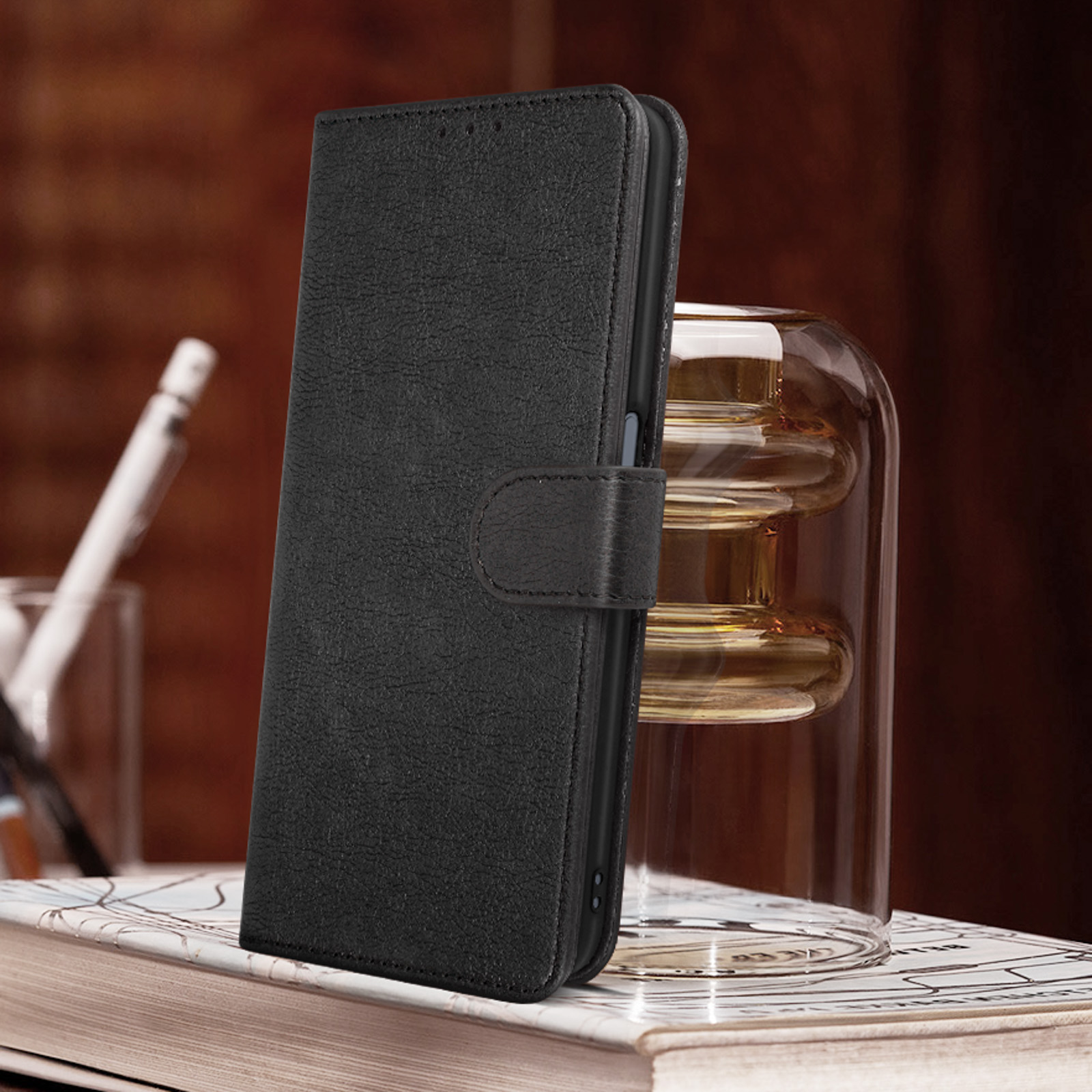 Una custodia per telefono in pelle nera con chiusura è appoggiata su un libro accanto a un vaso di vetro.