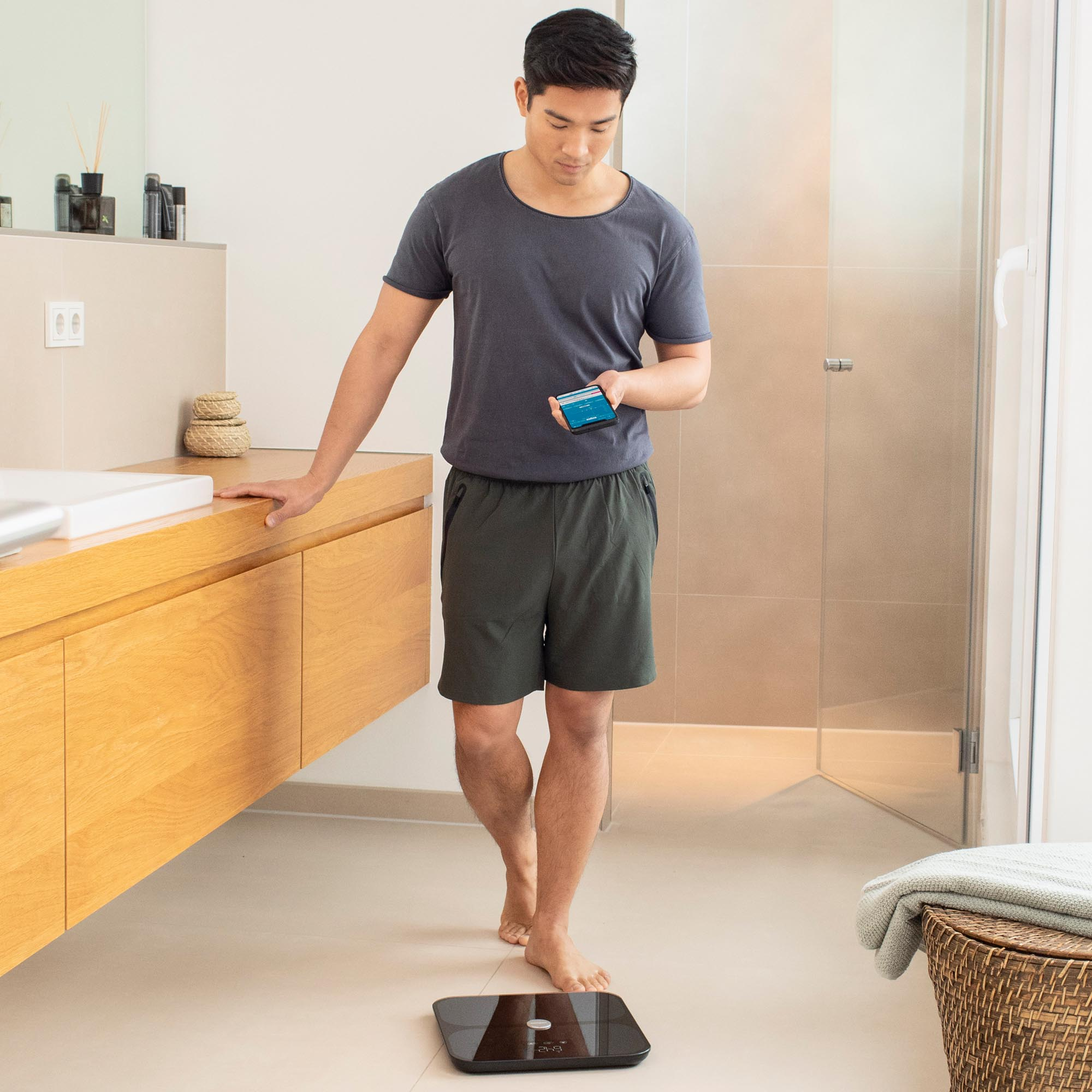 Uomo che usa una bilancia in bagno, tenendo un telefono. Dettagli della bilancia e del bagno.