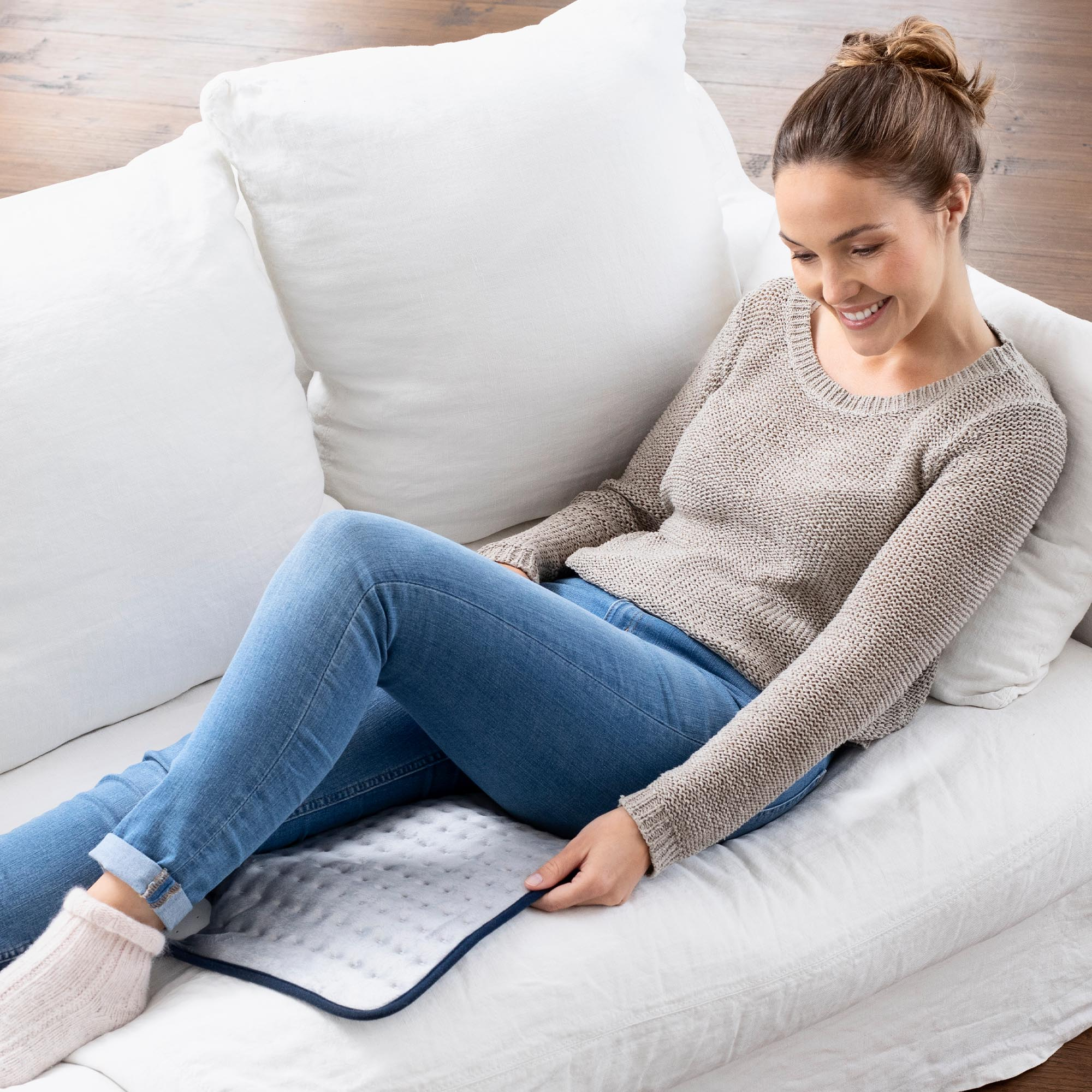 Kobieta na białej sofie z poduszką grzewczą, w dżinsach i swetrze.