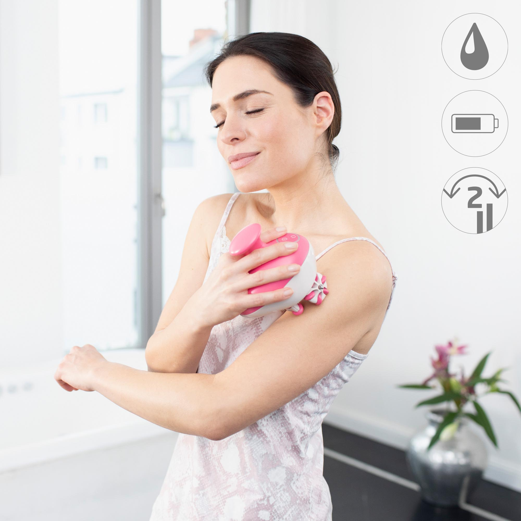 A woman using a pink and white facial massager on her shoulder. She is closing her eyes.