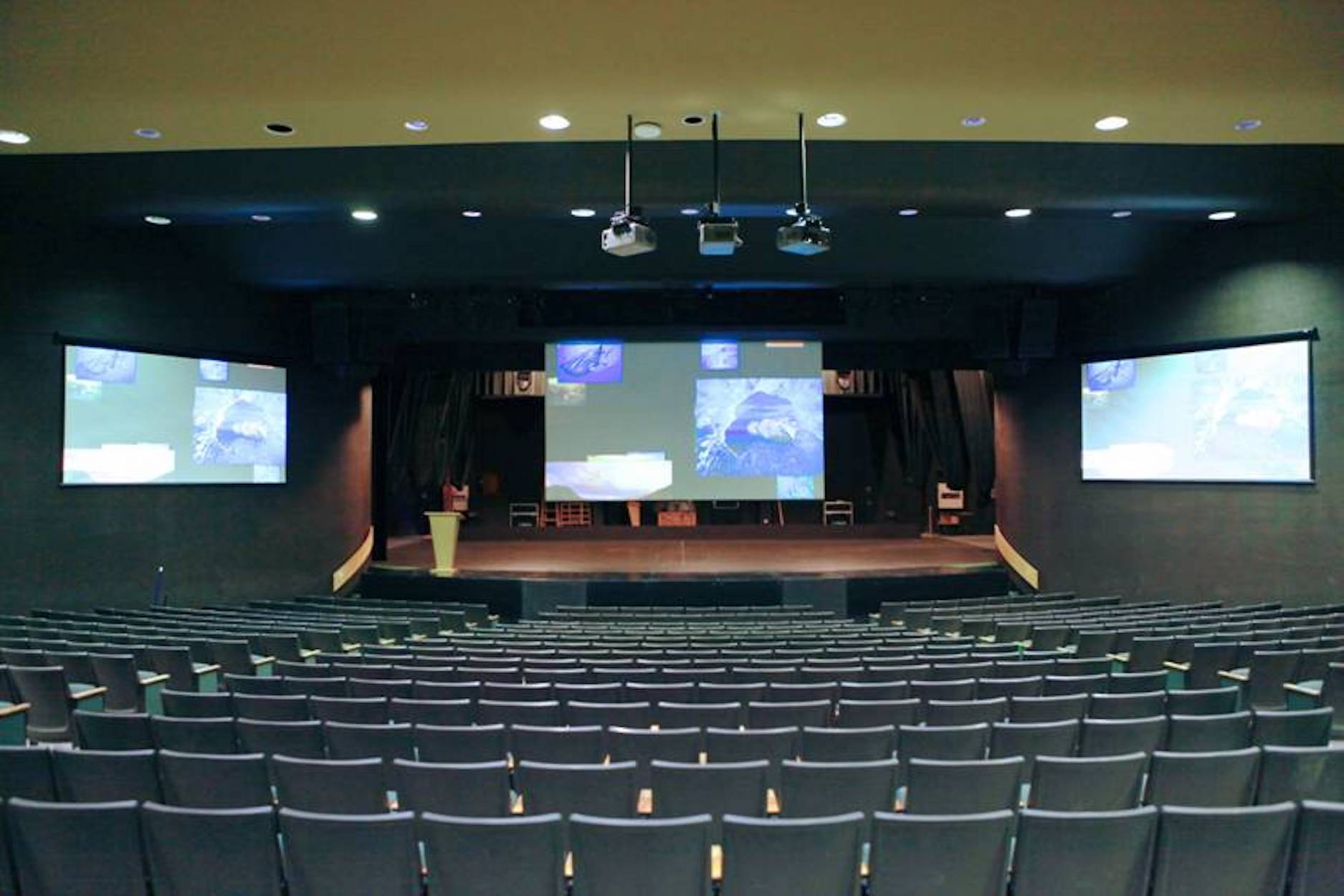 Auditorium with rows of seats. Two screens on the walls. Stage in the middle. Projectors on the ceiling.