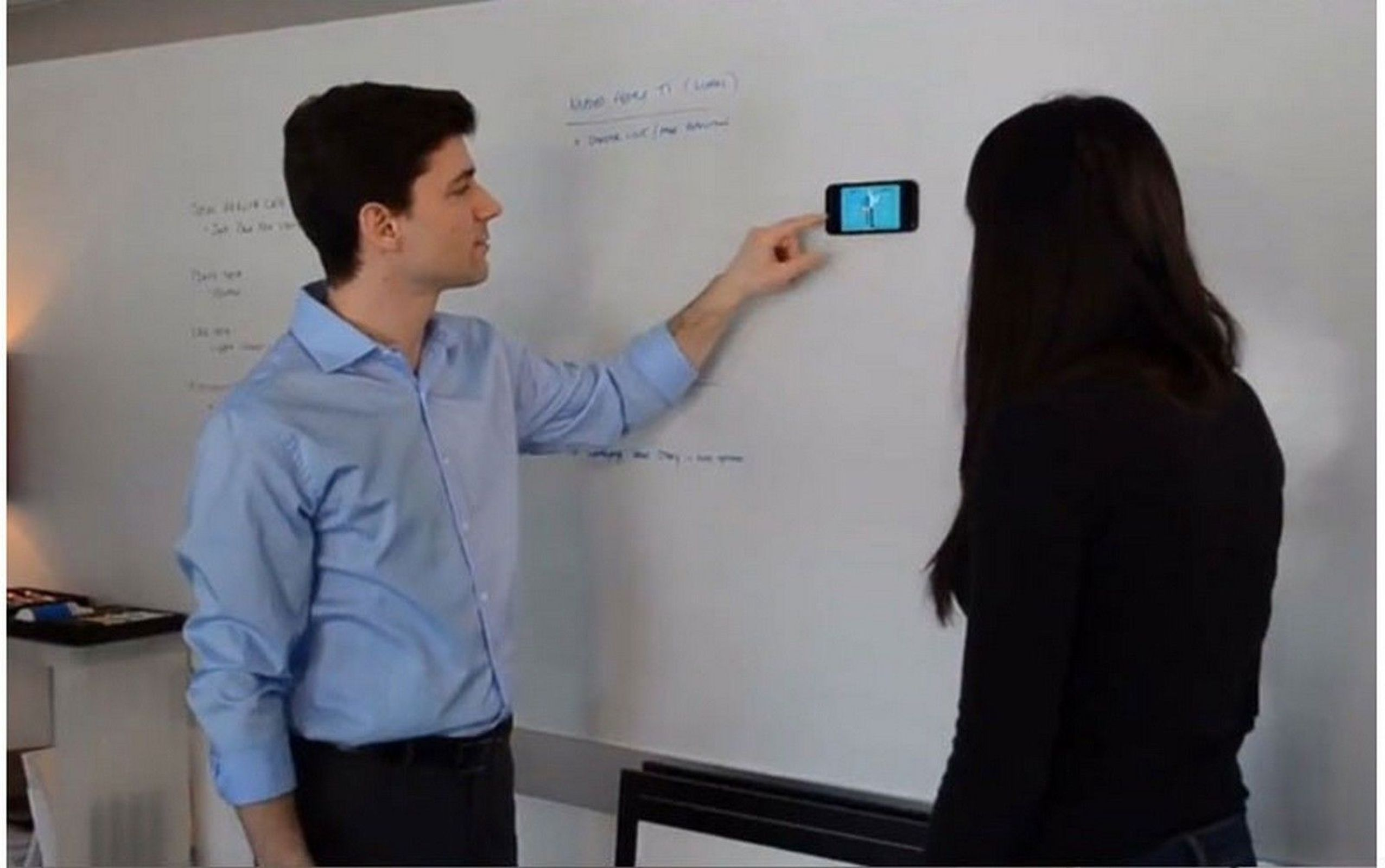 Mann zeigt auf Telefon an der Wand, Frau schaut es an. Whiteboard mit Schrift im Hintergrund.