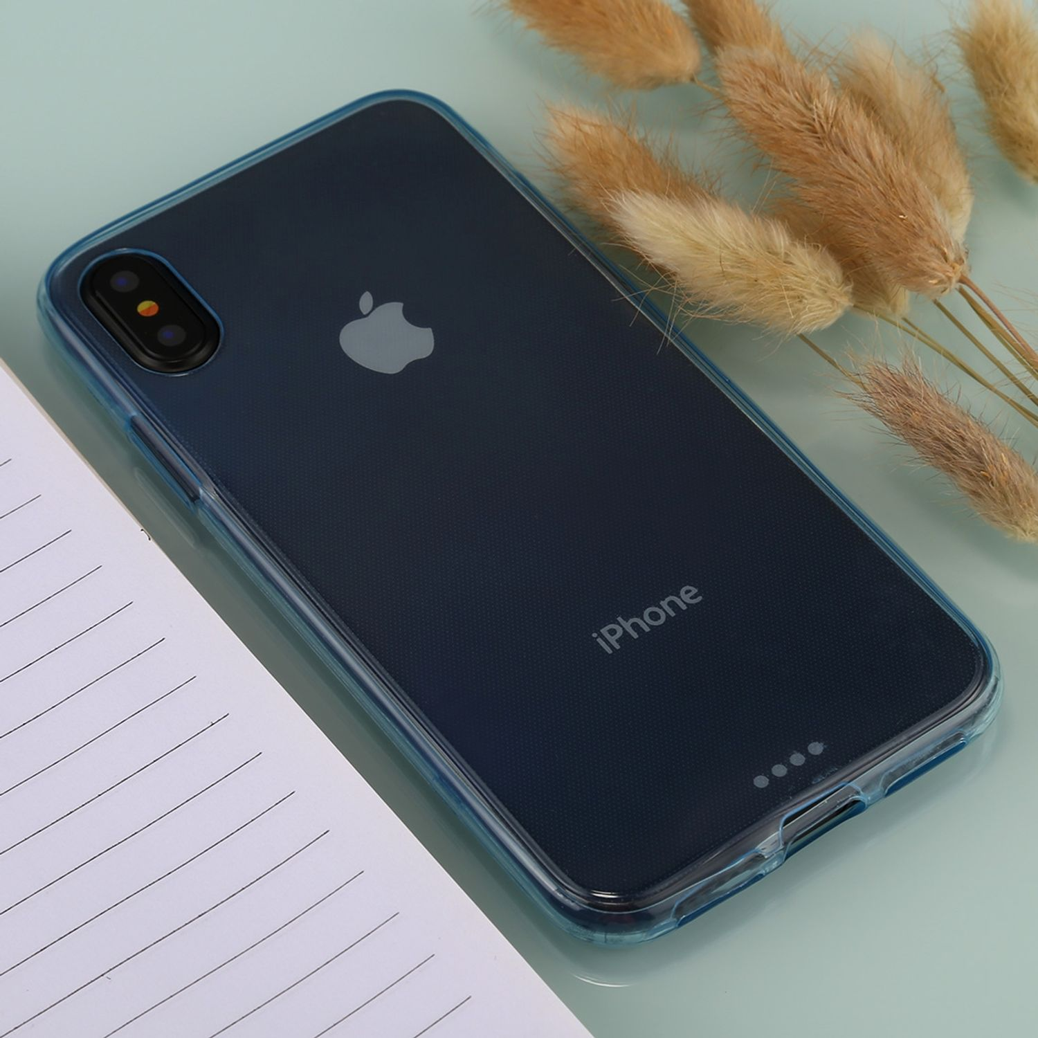 Transparente iPhone-Hülle auf einem Tisch neben einem Notizbuch und getrockneten Pflanzen.