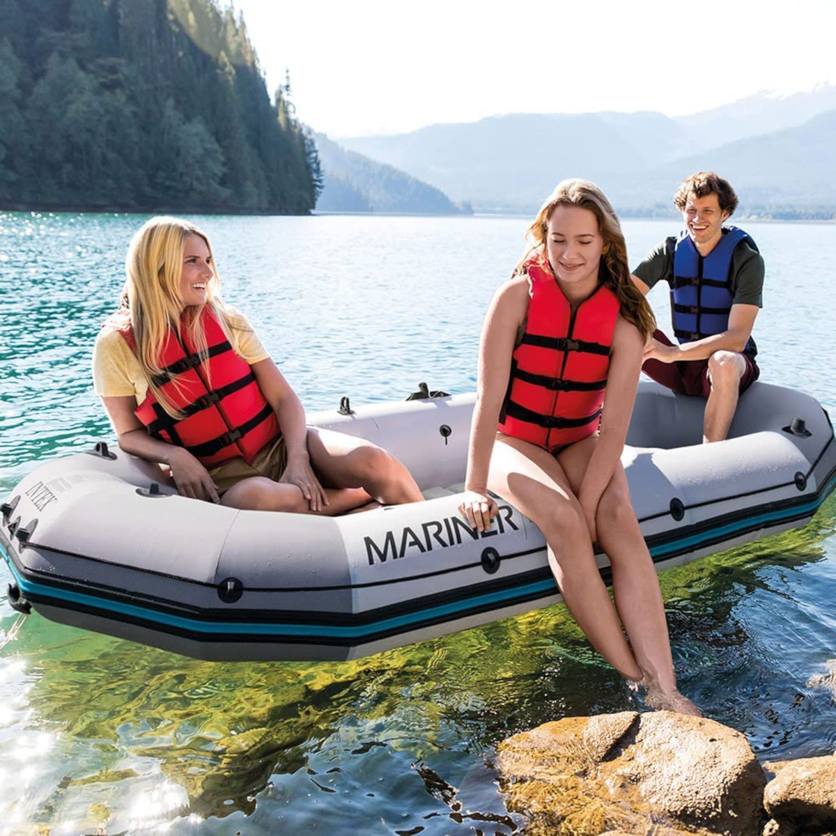 Drei Personen in einem grauen Boot auf einem See. Zwei haben rote Schwimmwesten. Berge im Hintergrund.