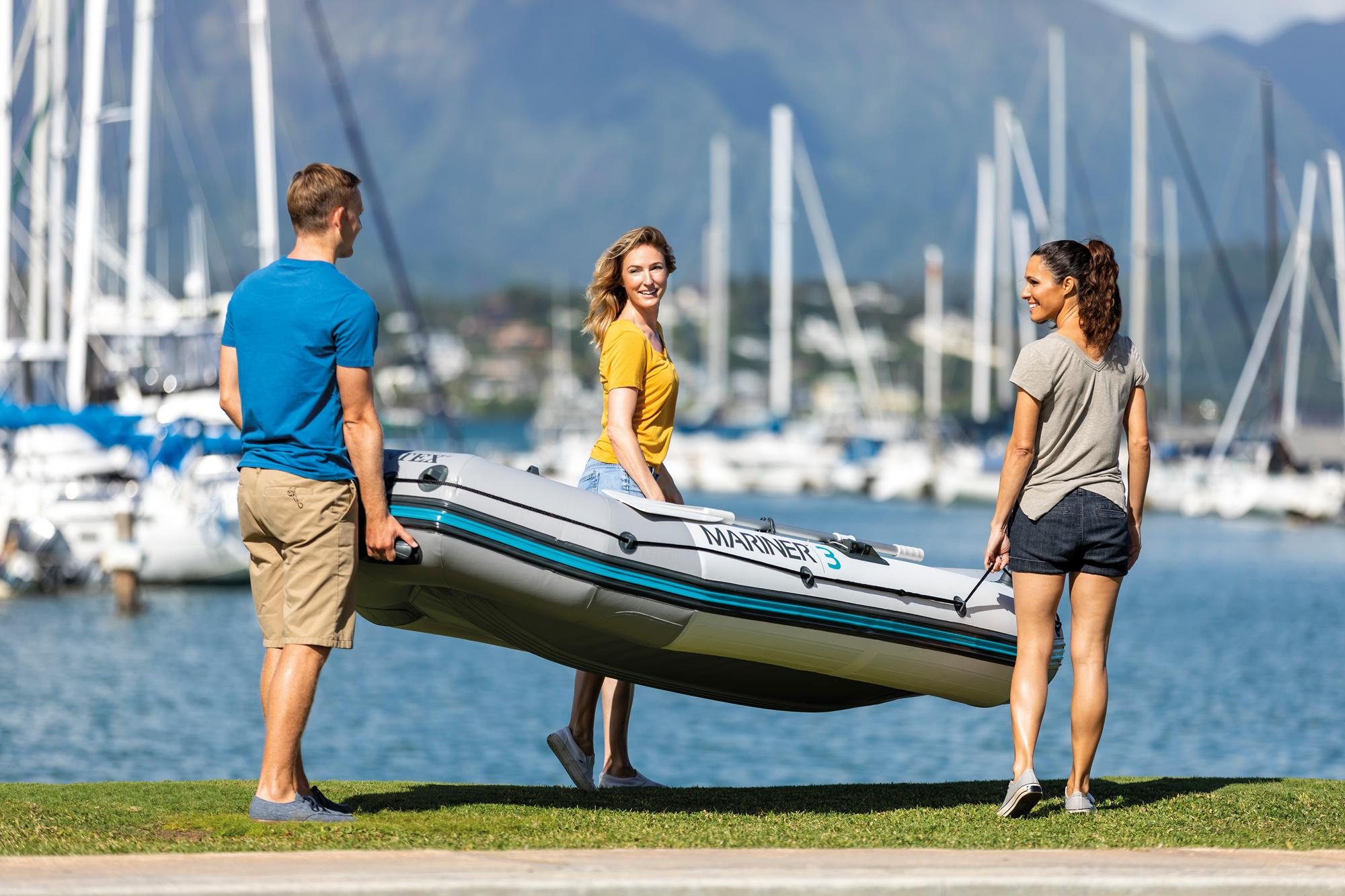 Drei Personen tragen ein graues und blaues Schlauchboot auf einer Wiese in der Nähe eines Sees.