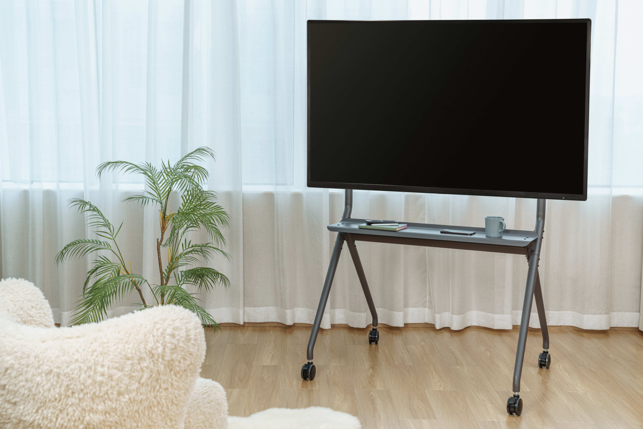 Ein großer schwarzer Fernseher steht auf einem rollenden grauen Ständer. Im Hintergrund sind eine Pflanze, ein Stuhl und Vorhänge.