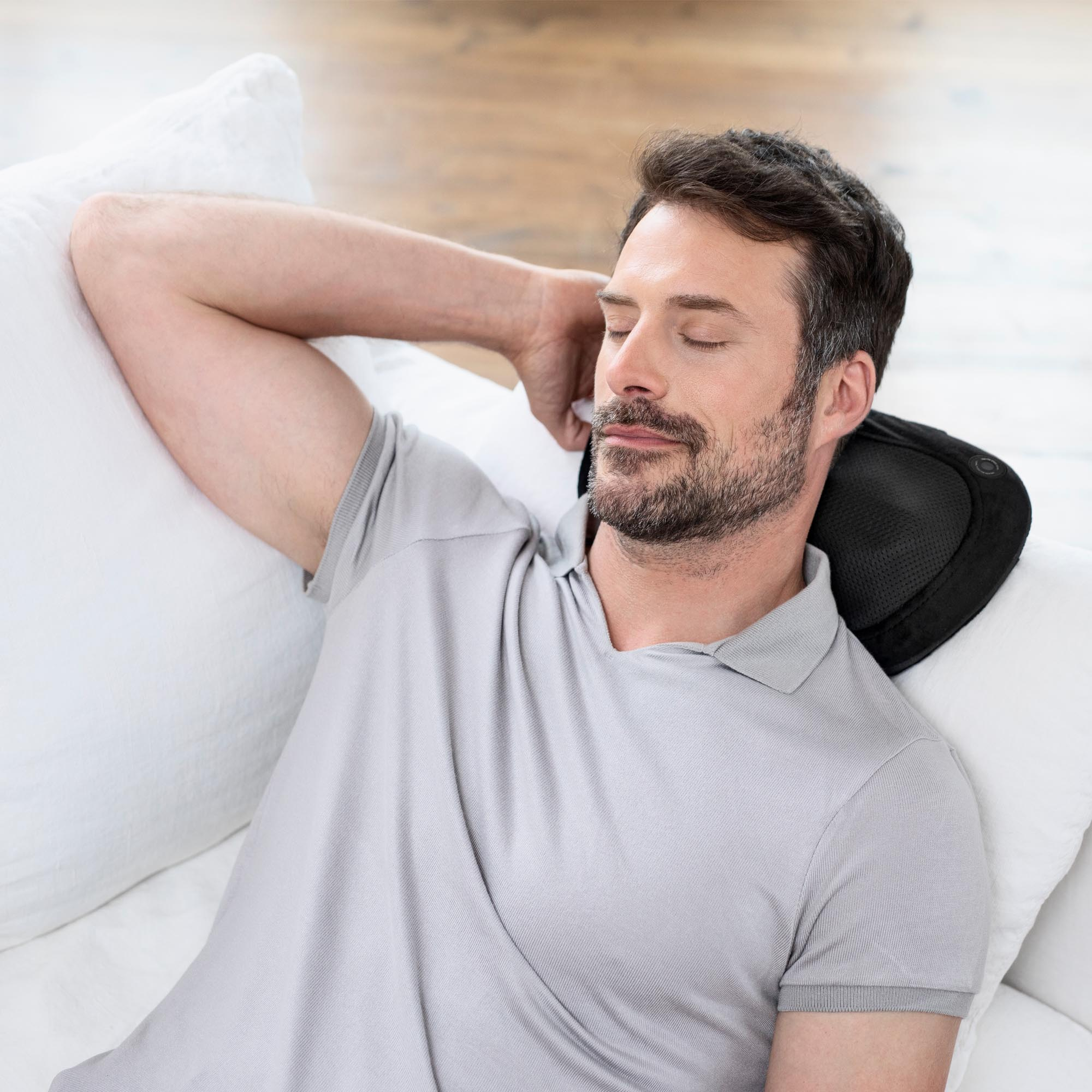 Mann entspannt sich auf weißem Sofa mit schwarzem Nackenmassagekissen.