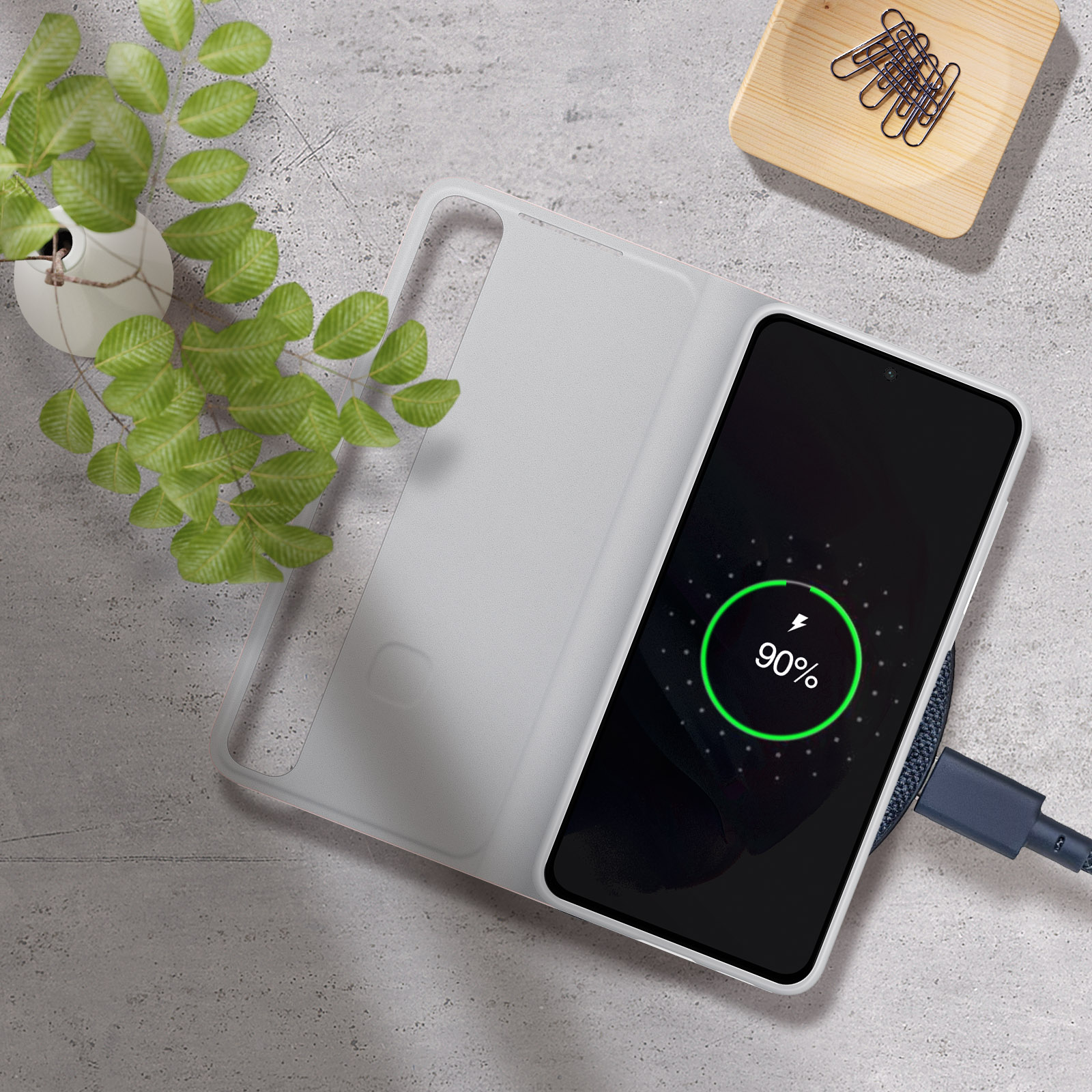 Telefon lädt auf einem Pad. Ladeanzeige 90% auf dem Bildschirm. Pflanze und Papiere.