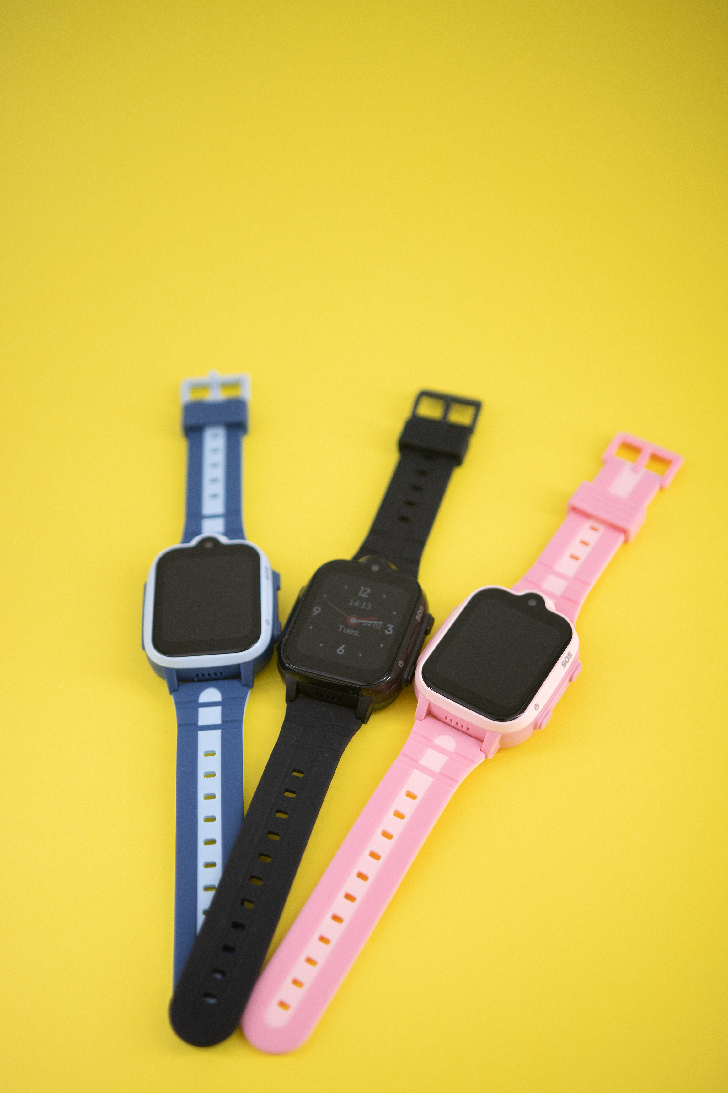 Tres smartwatches con correas de diferentes colores: azul, negro y rosa. Fondo amarillo.