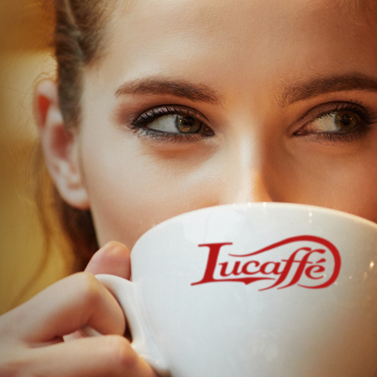 Frau hält eine weiße Tasse mit 'Lucaffe'-Logo. Nahaufnahme ihres Gesichts, Augen schauen weg.