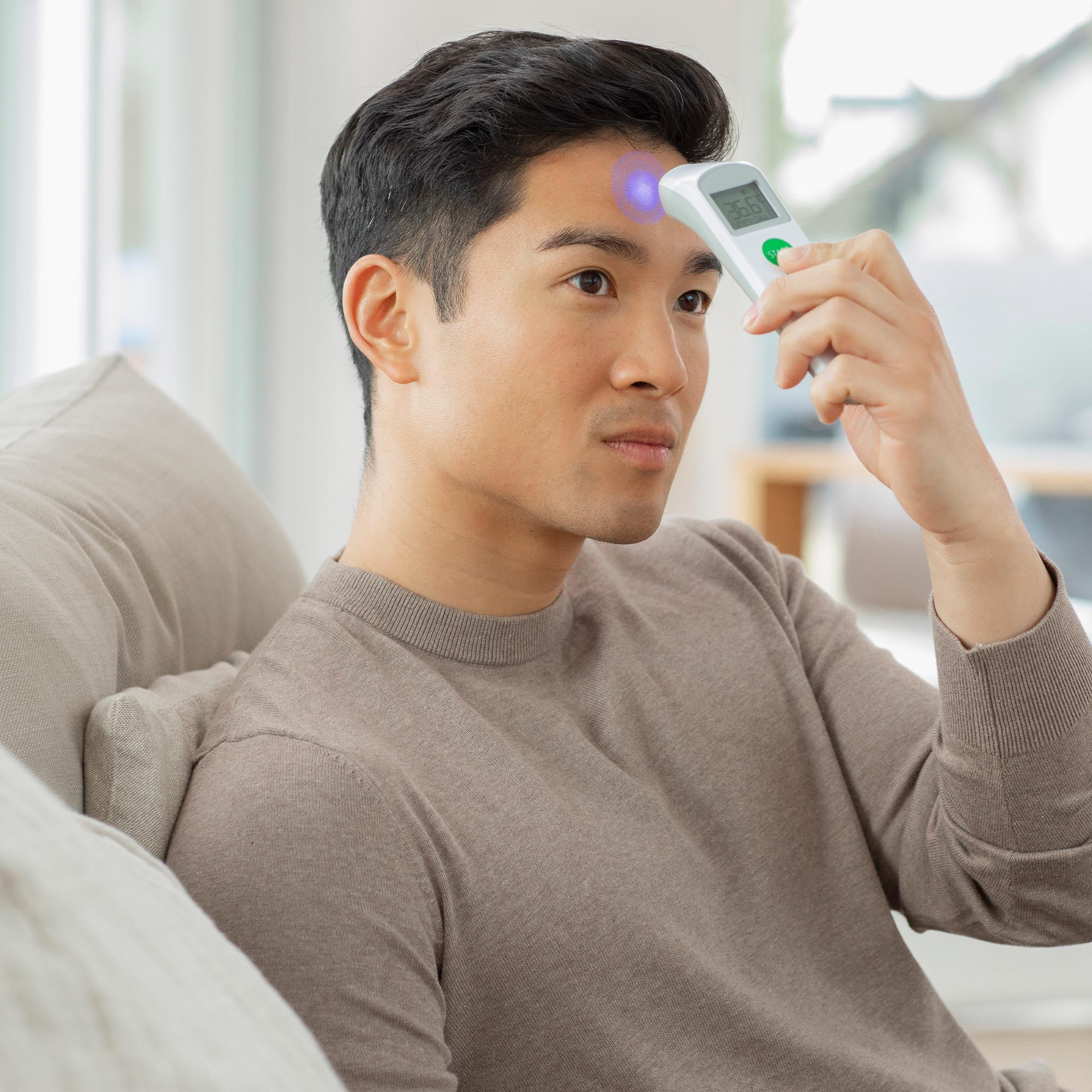 Mężczyzna używa termometru cyfrowego na czole. Siedzi na kanapie.