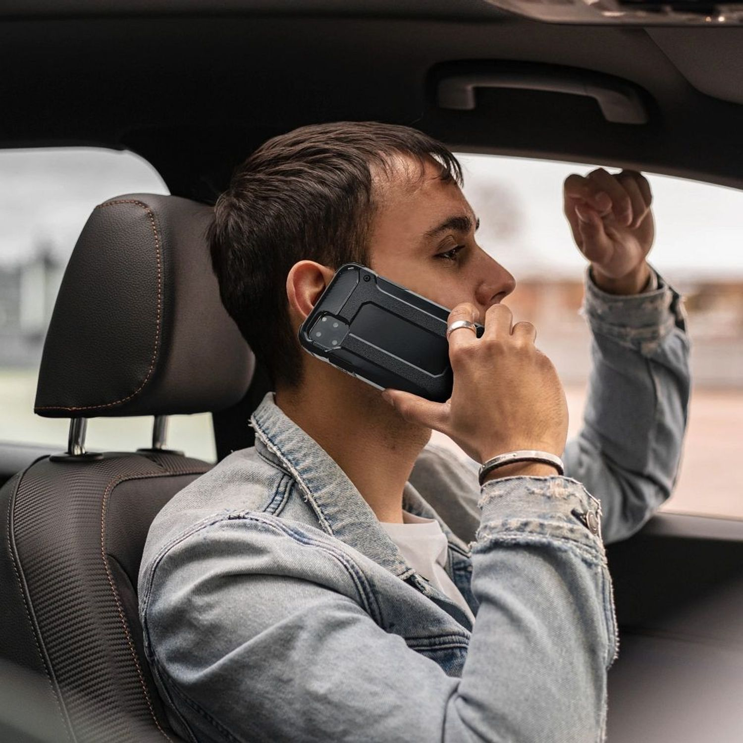 Mann in Jeansjacke telefoniert mit schwarzem Handy im Auto. Er hält das Telefon mit einer Hülle.