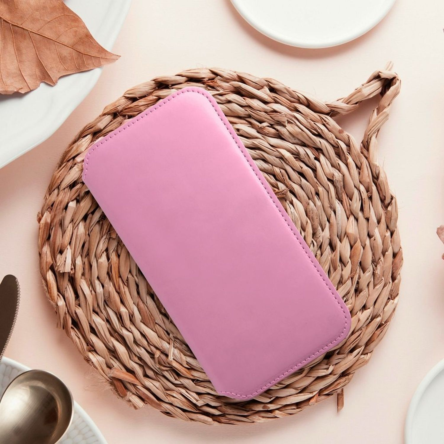 Rosa Handyhülle auf einer gewebten Matte mit Blatt und Tellern im Hintergrund.