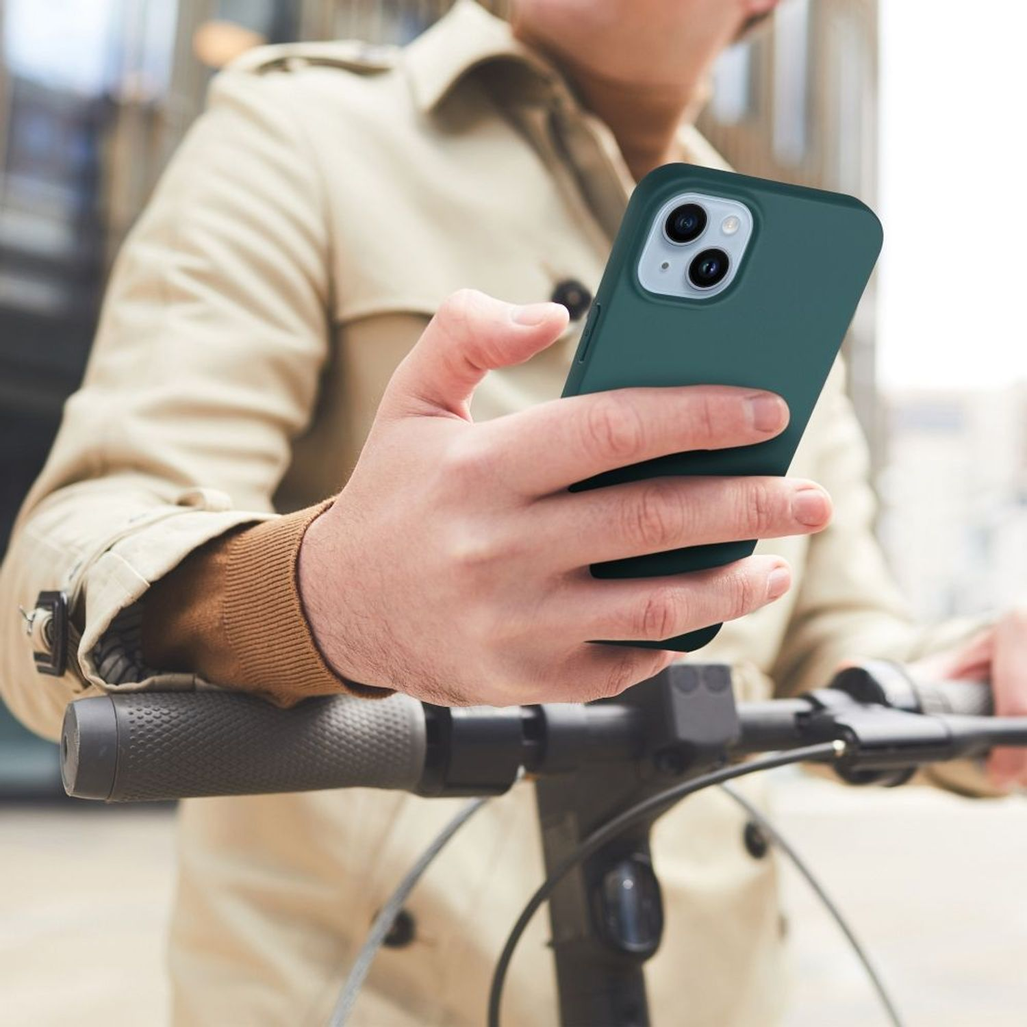 Person hält grünes Telefon, auf einem Elektroroller, in städtischer Umgebung. Beiger Mantel getragen.