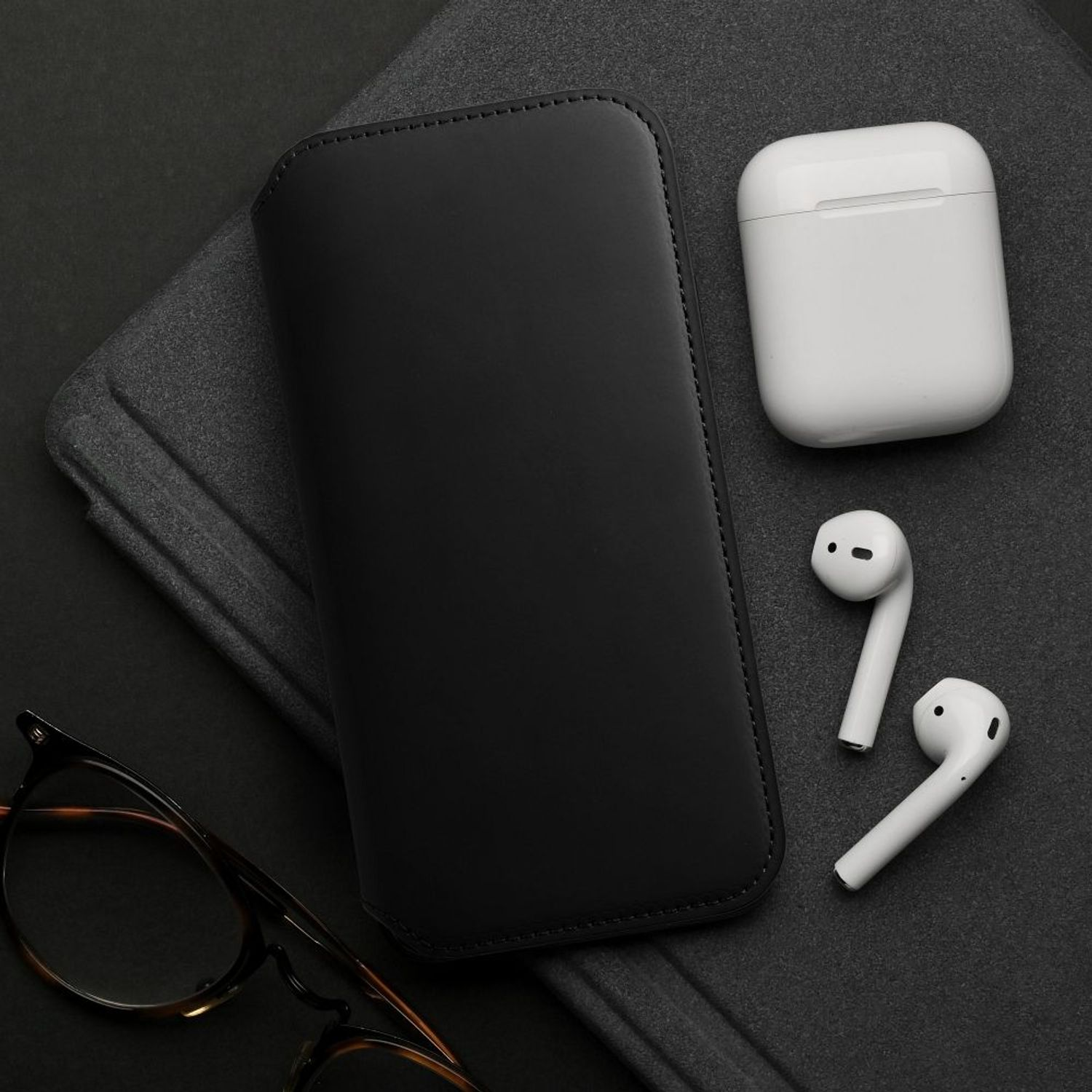 Schwarze Leder-Handyhülle, AirPods und Brille auf dunkelgrauer Oberfläche. Gut beleuchtet.