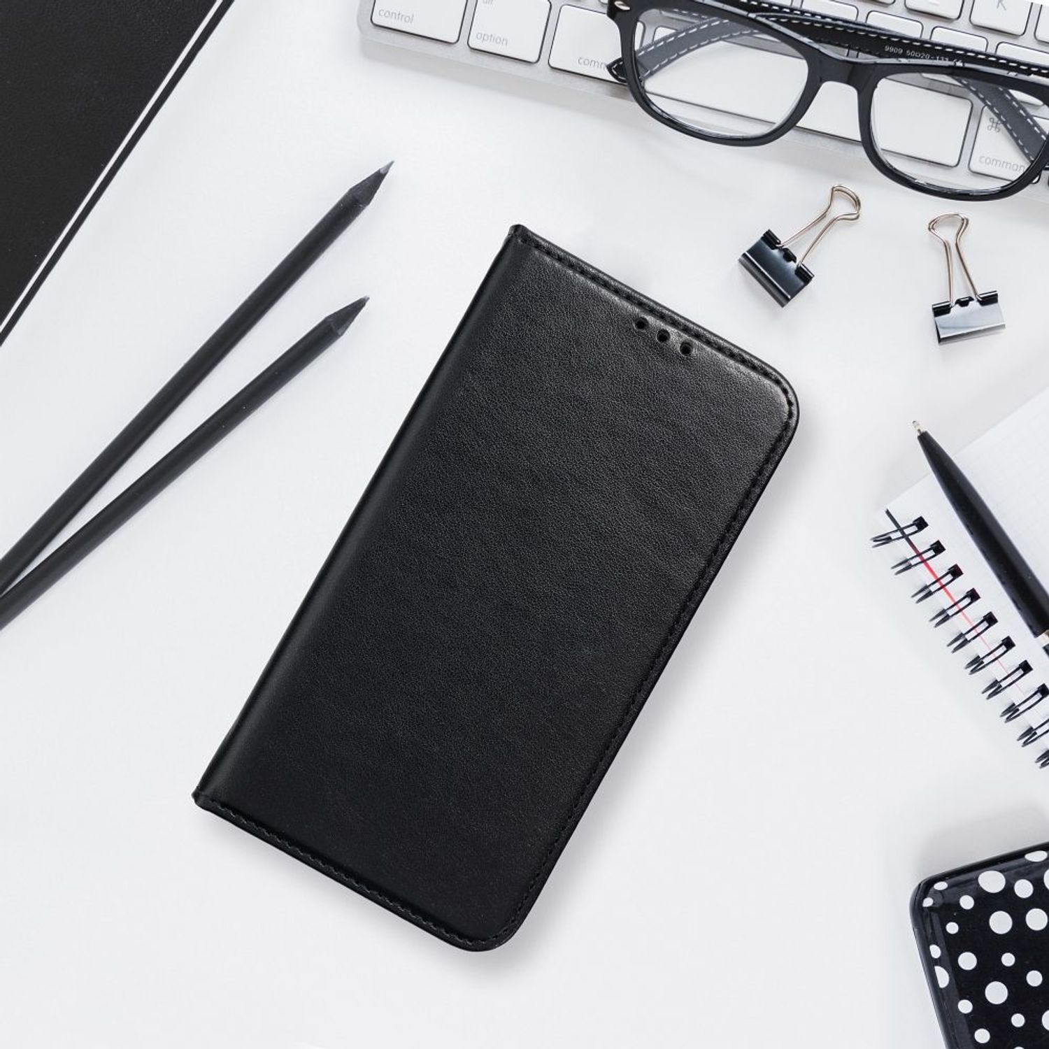Schwarze Handyhülle mit Stift, Brille und Notizblock auf weißem Schreibtisch.