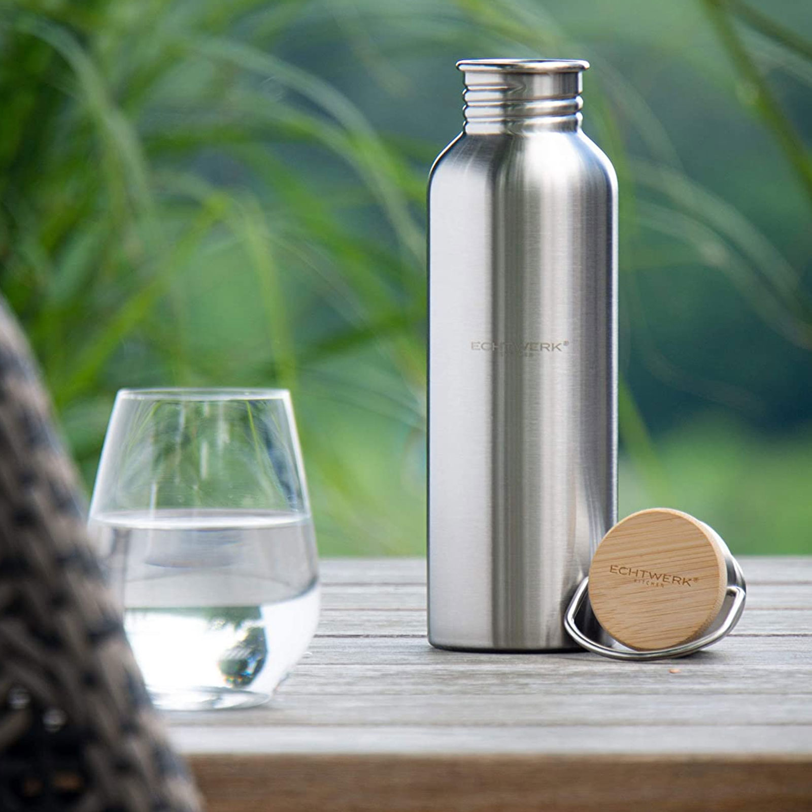 Eine Edelstahl-Wasserflasche, ein Glas mit Wasser und ein Holzdeckel auf einem Holztisch.