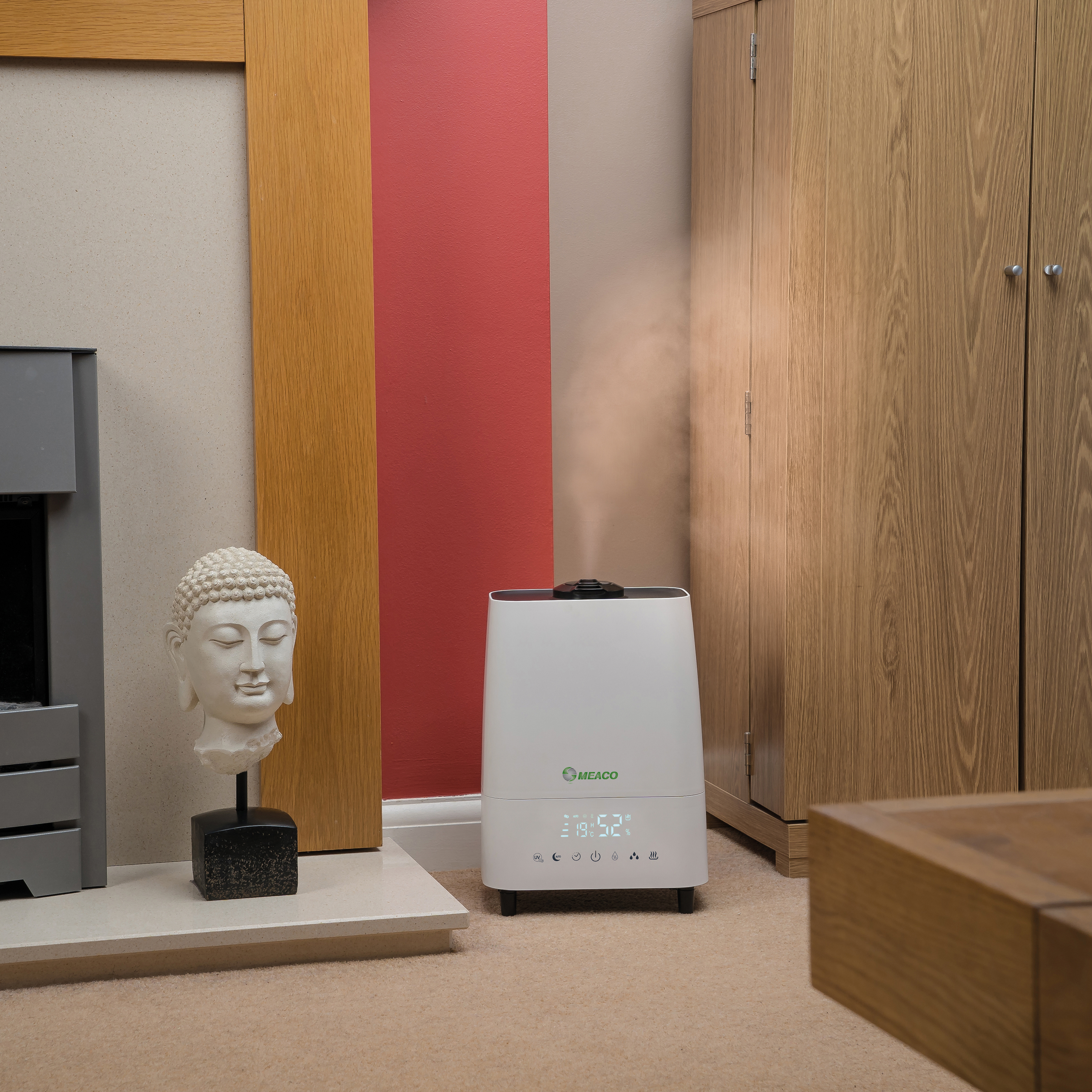 Biały nawilżacz powietrza w pokoju. Obok znajduje się posąg głowy Buddy i szafka. Pokój ma beżową wykładzinę.