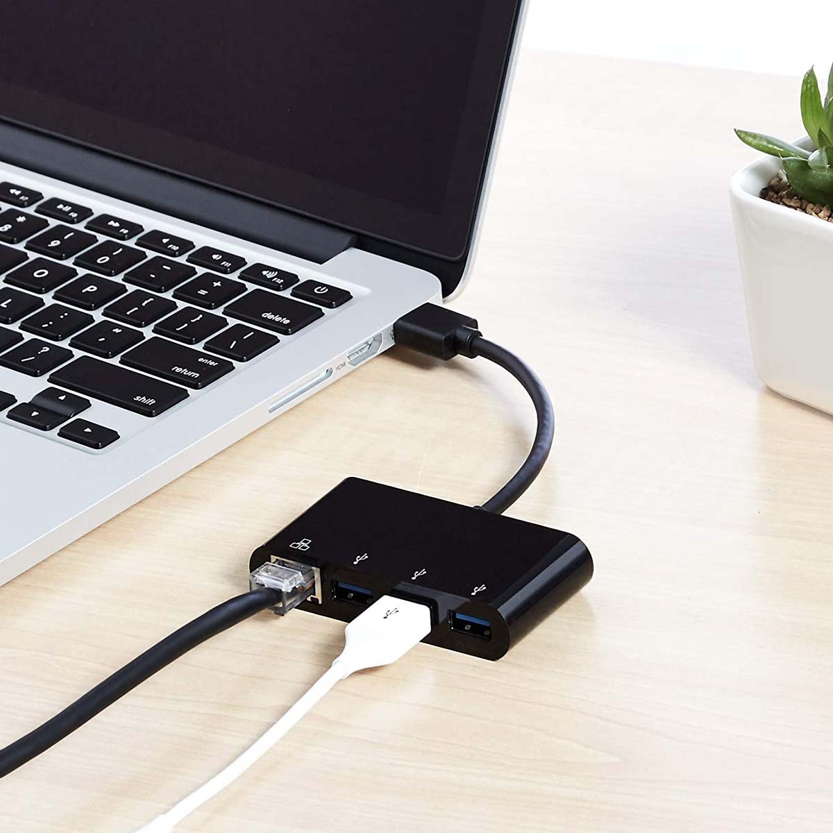 Laptop collegato a un hub nero con più porte. Cavo bianco e pianta sulla scrivania.