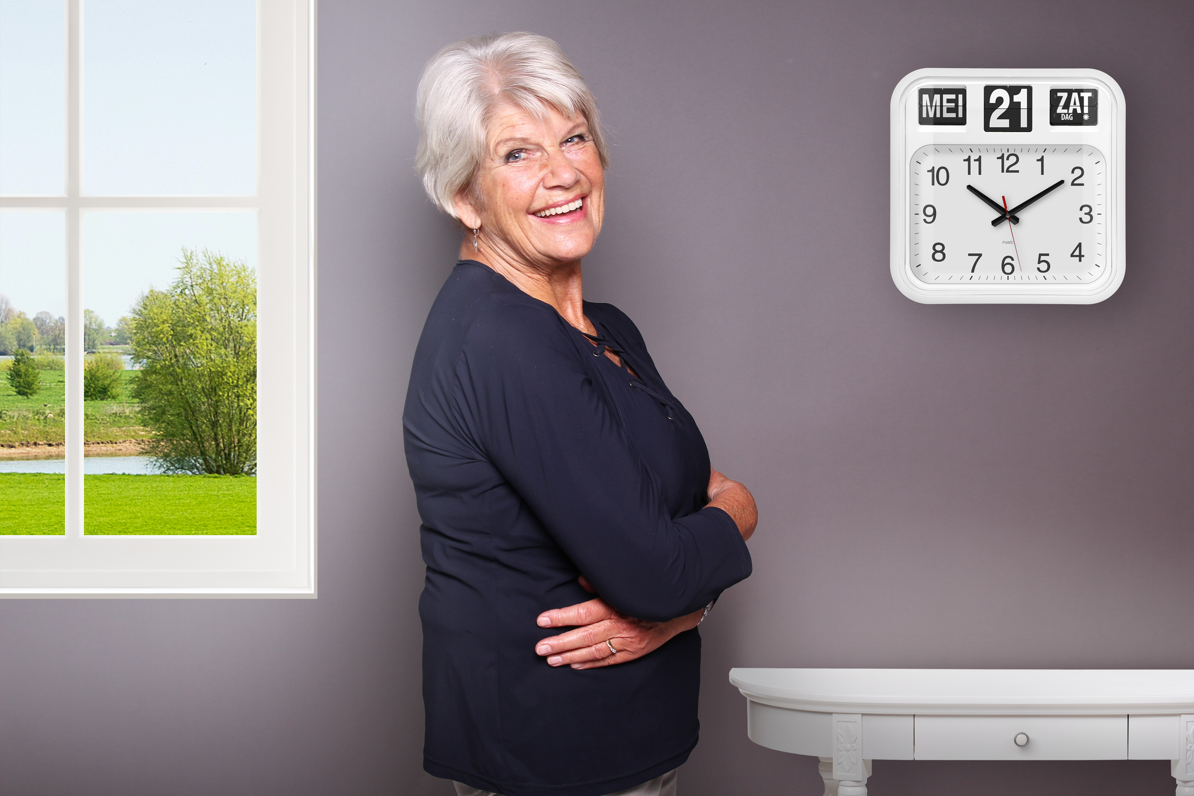 Eine ältere Frau lächelt, trägt ein dunkelblaues Oberteil, steht neben einer weißen Wand mit einer Uhr.