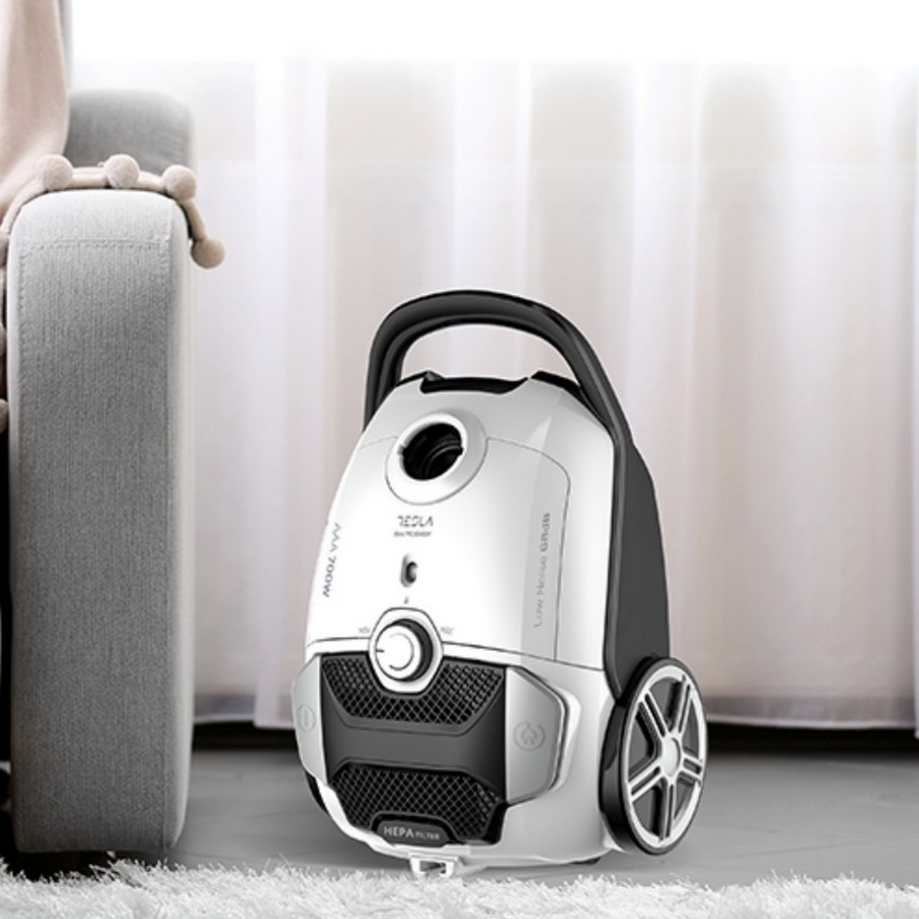 Biało-czarny odkurzacz stoi na białym dywanie, obok szarej sofy.