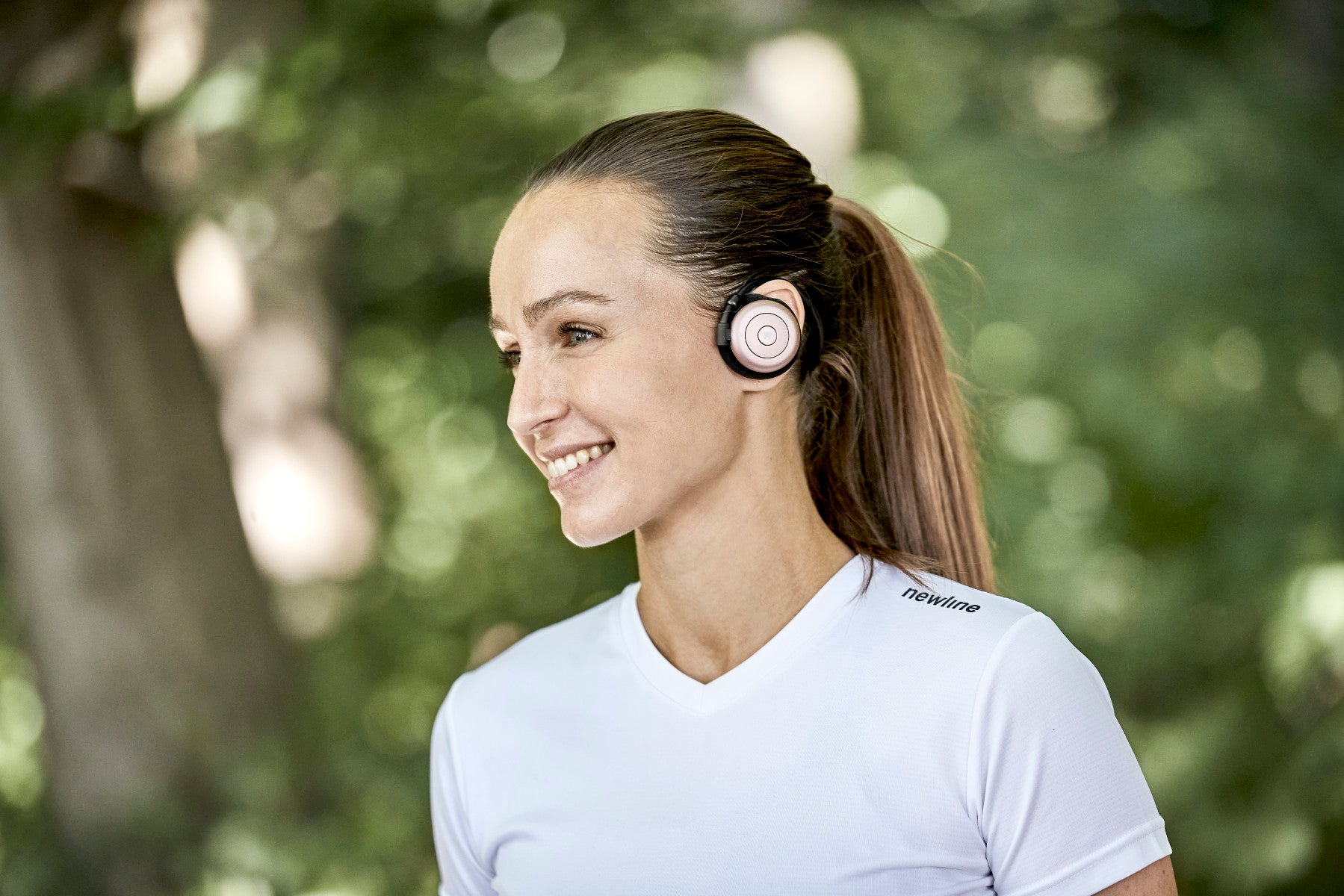 Mujer sonriendo, usando auriculares inalámbricos rosas y negros. Lleva una camiseta blanca.