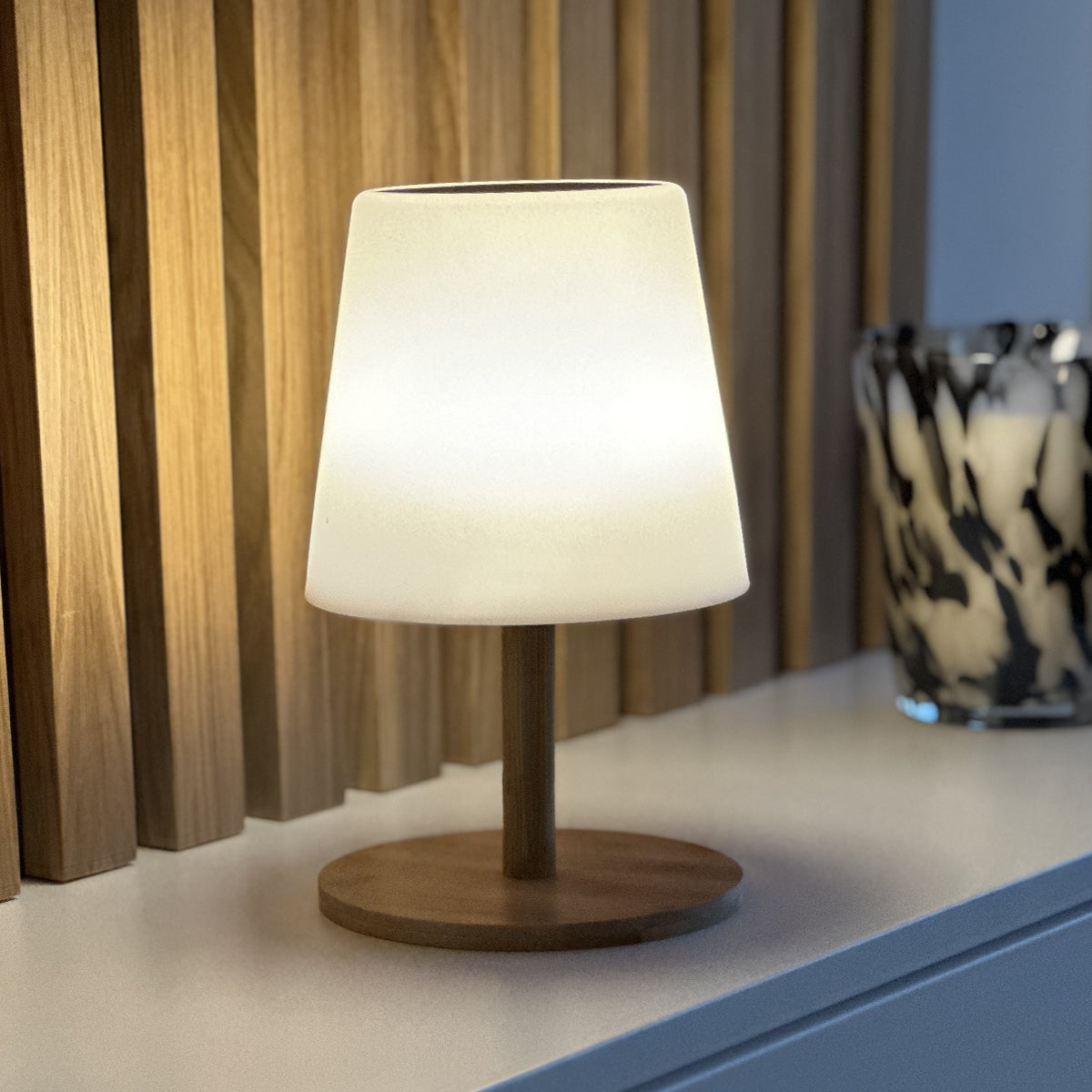Eine kleine, weiße Lampe auf einem Holzständer, platziert auf einer weißen Oberfläche. Rechts ein Glas.