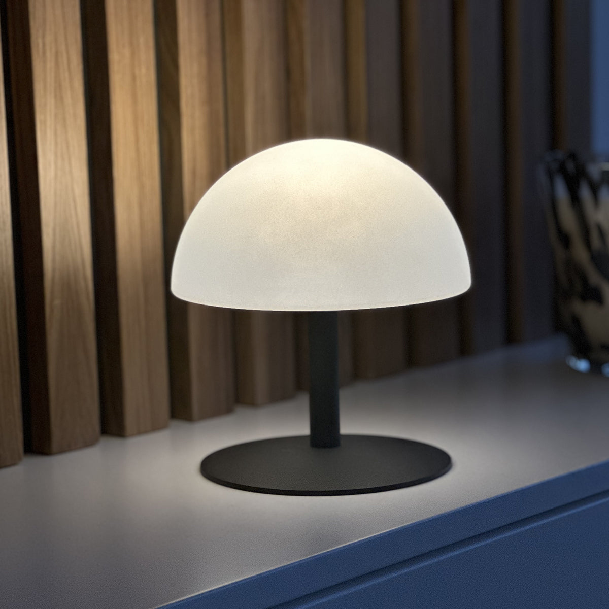 Eine moderne Tischlampe mit einem weißen, halbkugelförmigen Schirm und einem schwarzen Sockel auf weißer Oberfläche.