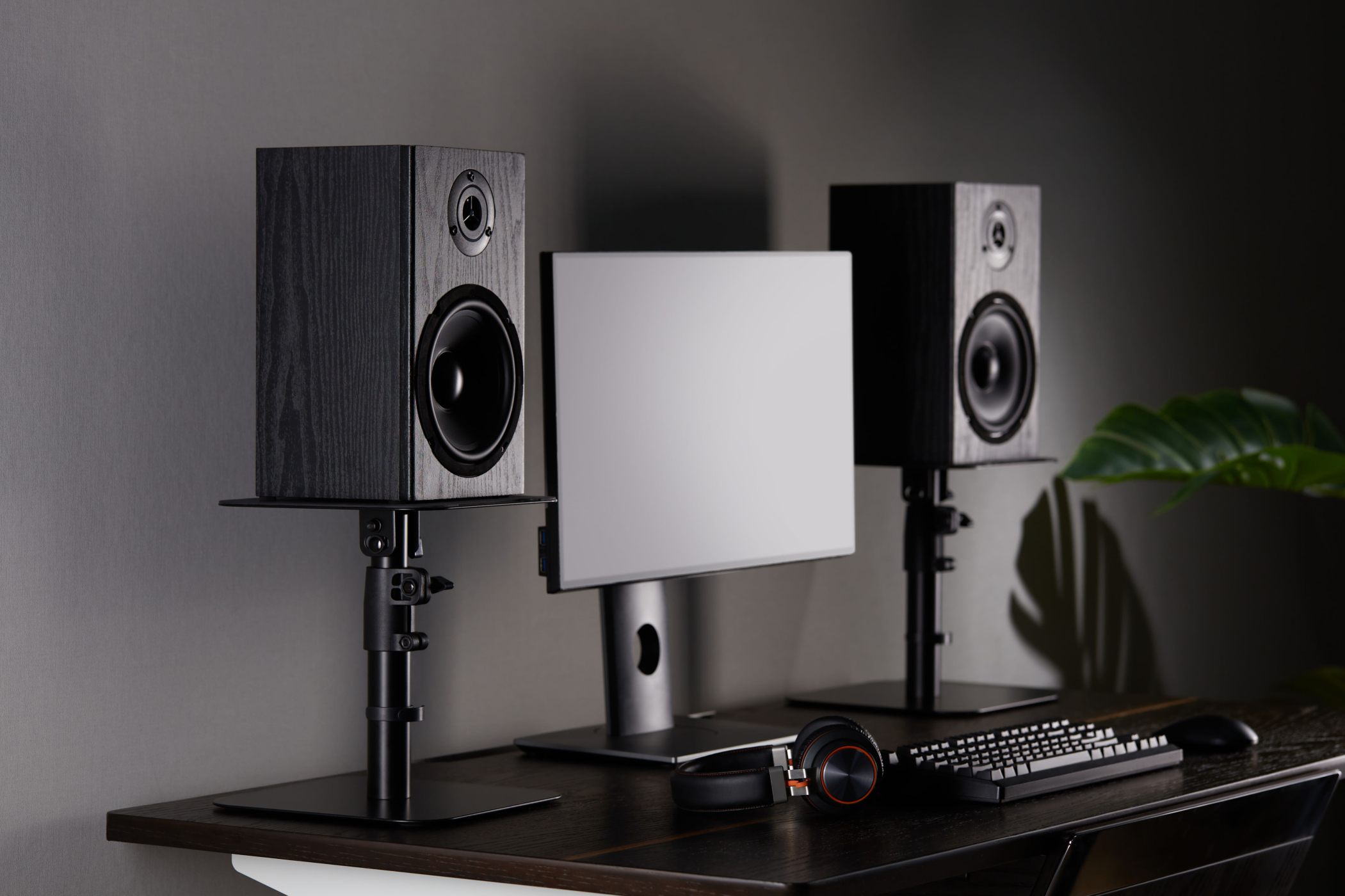 Two black speakers on stands flanking a monitor, with a keyboard and headphones on a desk.