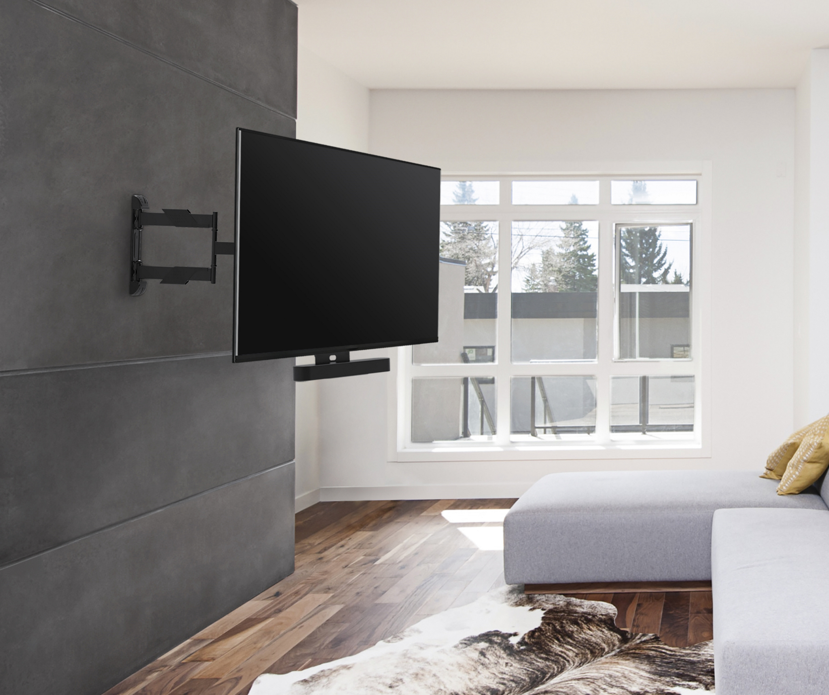 A large TV mounted on a dark wall, with a soundbar below. A window and a sofa are visible.