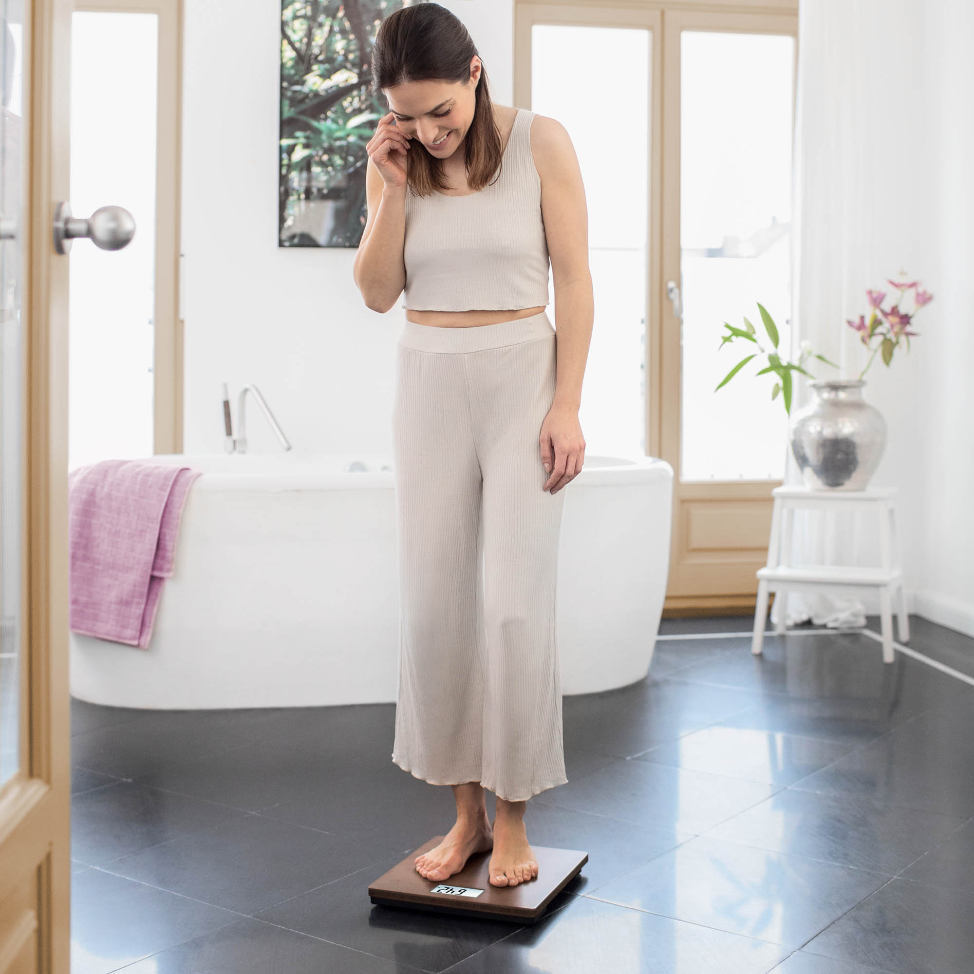 Mujer de pie sobre una báscula digital en un baño, sonriendo. Hay una bañera y una planta en el fondo.