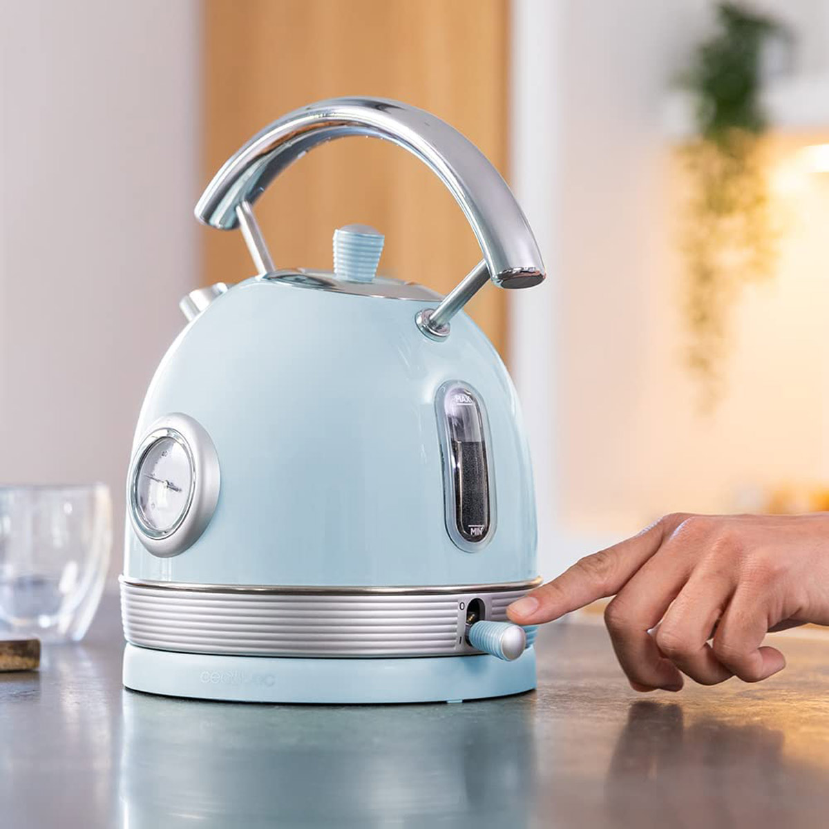 Jasnoniebieski czajnik z chromowanym uchwytem i wskaźnikiem poziomu wody. Ręka naciska przycisk.