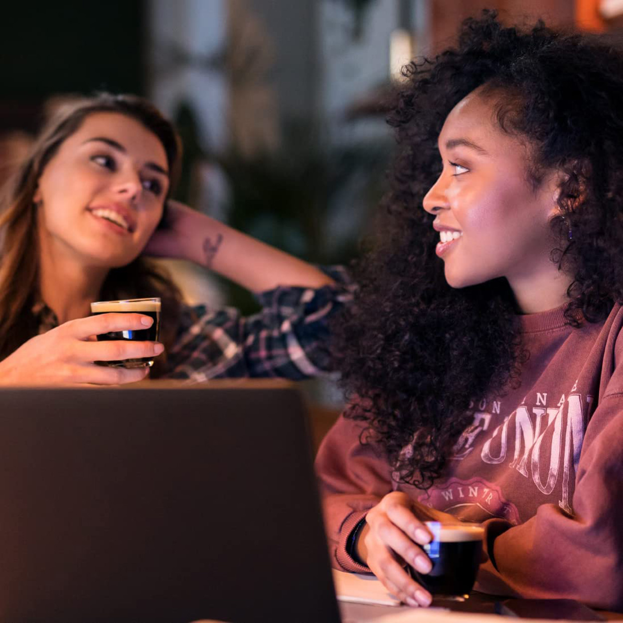 Zwei Frauen sitzen in einem Café, eine hält ein Getränk, eine benutzt einen Laptop.
