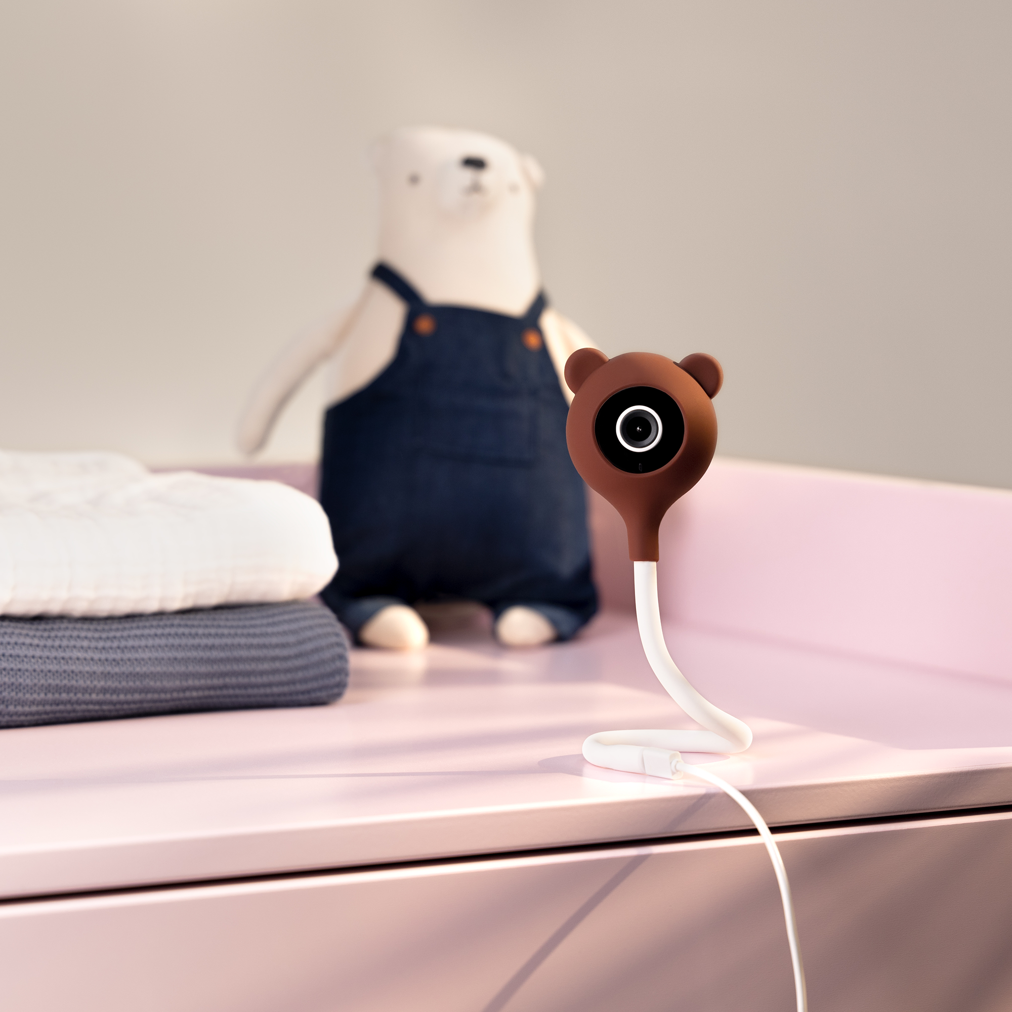 Ein Babyphone mit einer bärenförmigen Kamera und einem Plüschbären im Hintergrund auf rosa Oberfläche.