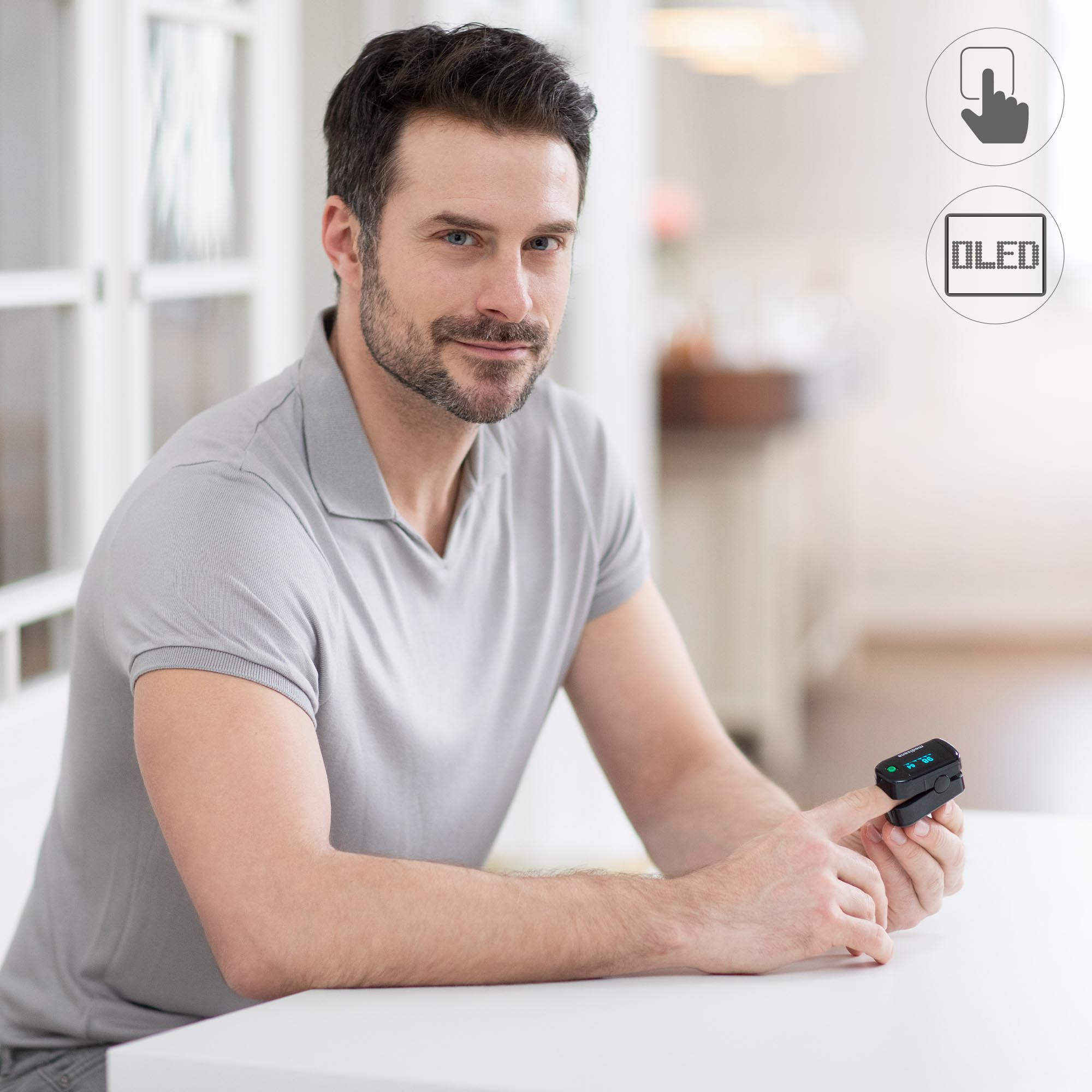 Mężczyzna ma na palcu pulsoksymetr. Siedzi przy białym stole.