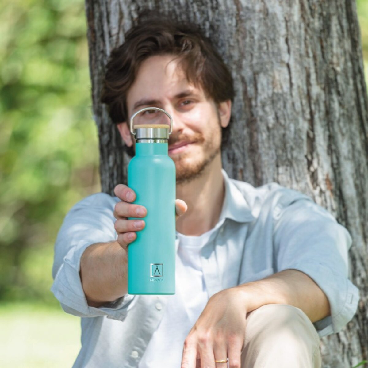 Un hombre sostiene una botella de agua verde azulado en un parque, sonriendo.