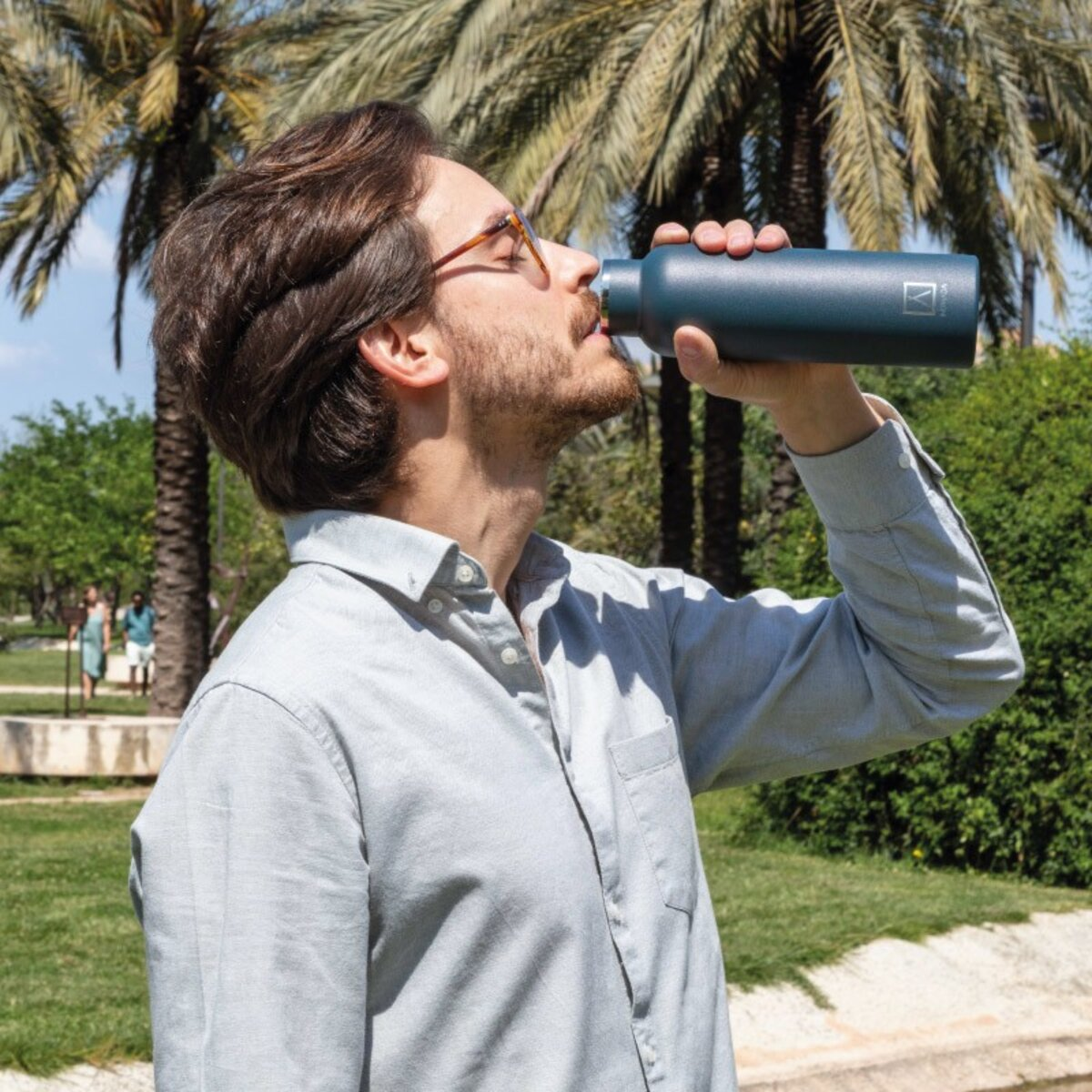 Hombre bebiendo de una botella de agua verde azulado, palmeras al fondo.