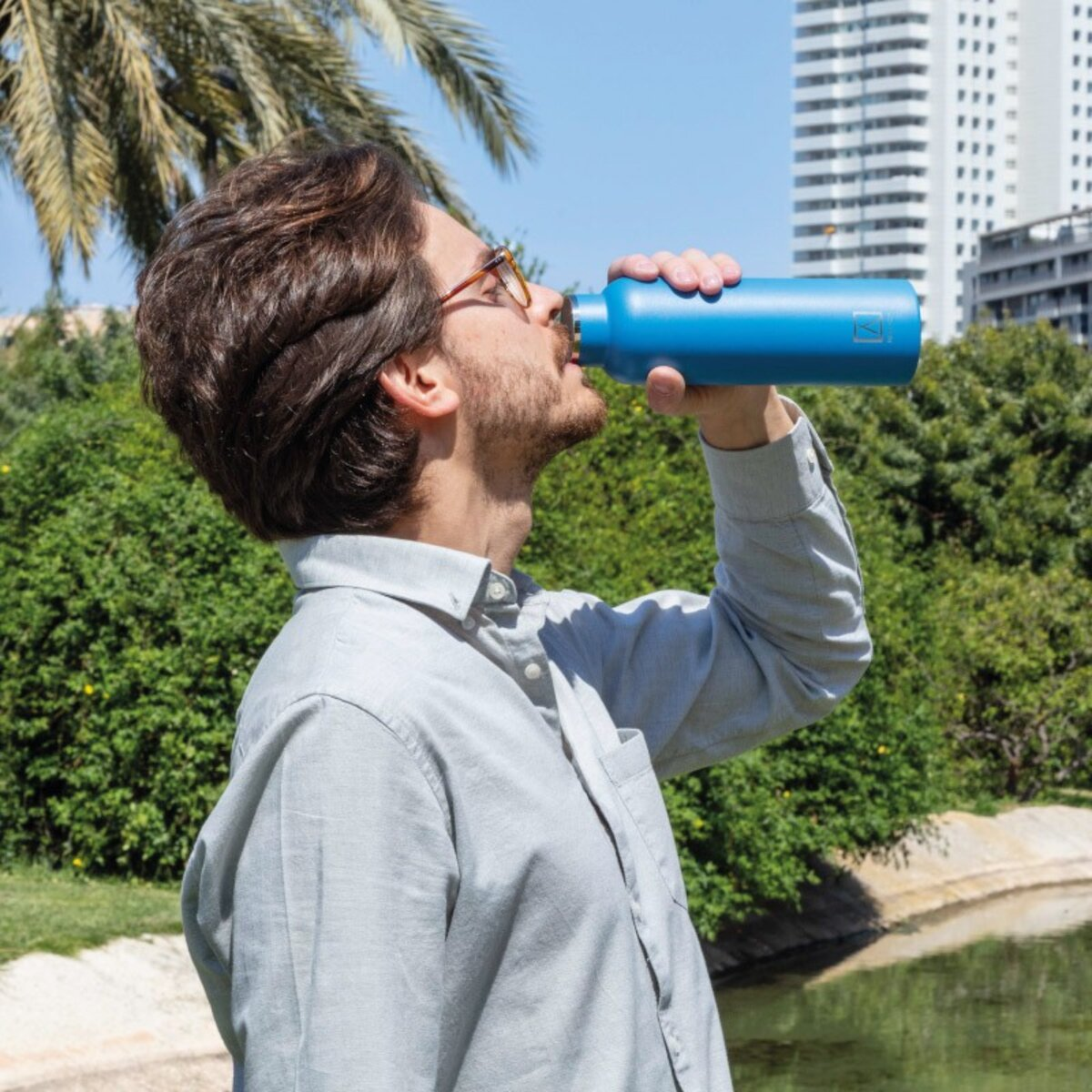 Hombre bebiendo de una botella de agua azul al aire libre. Follaje verde y lago al fondo.
