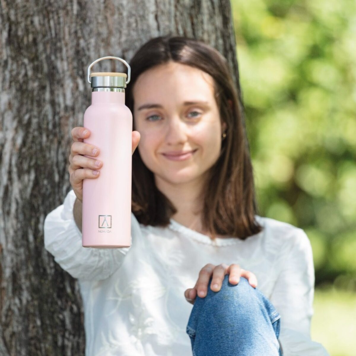 Mujer sostiene una botella de agua rosa frente a un árbol.