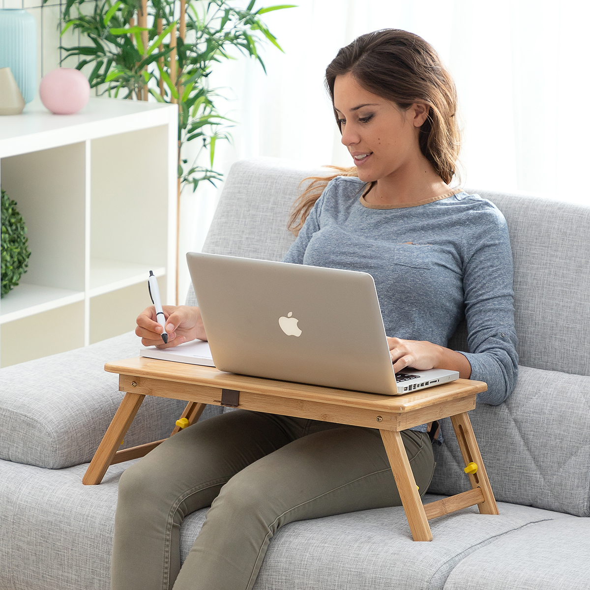 Donna che lavora su un laptop su un tavolino da grembo in legno, seduta su un divano grigio.