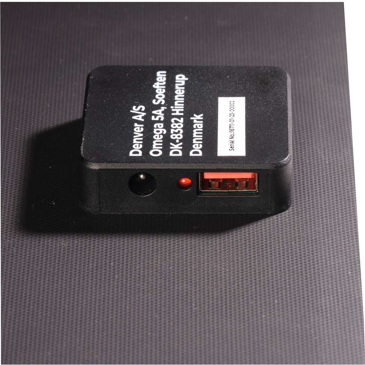 Czarna ładowarka z czerwonym światłem i portem USB. Tekst: Denver A/S, Omega 5A, DK-8382 Hinnerup Dania.