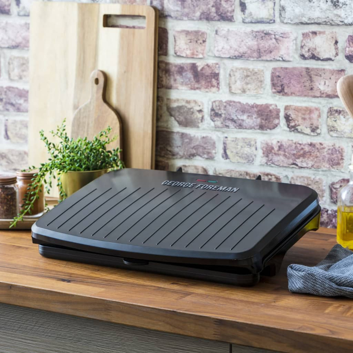 Ein George Foreman Grill auf einer Holzoberfläche, mit Ziegelmauer im Hintergrund und Küchenutensilien.