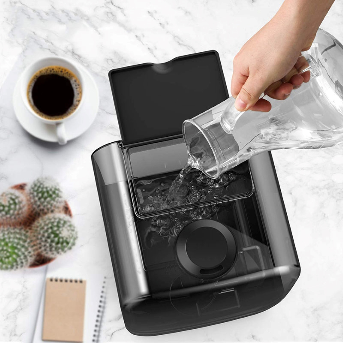 Eine Hand gießt Wasser in einen schwarzen Wassertank. Eine Tasse Kaffee und ein Kaktus sind sichtbar.